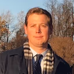Man with light brown hair wearing a dark jacket, striped scarf, and blue tie outdoors with trees in the background.