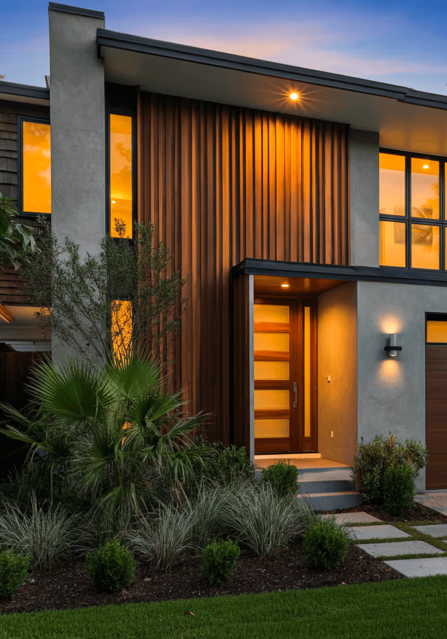 Modern house exterior at dusk with illuminated wooden front door, large vertical wood paneling, and landscaped greenery.