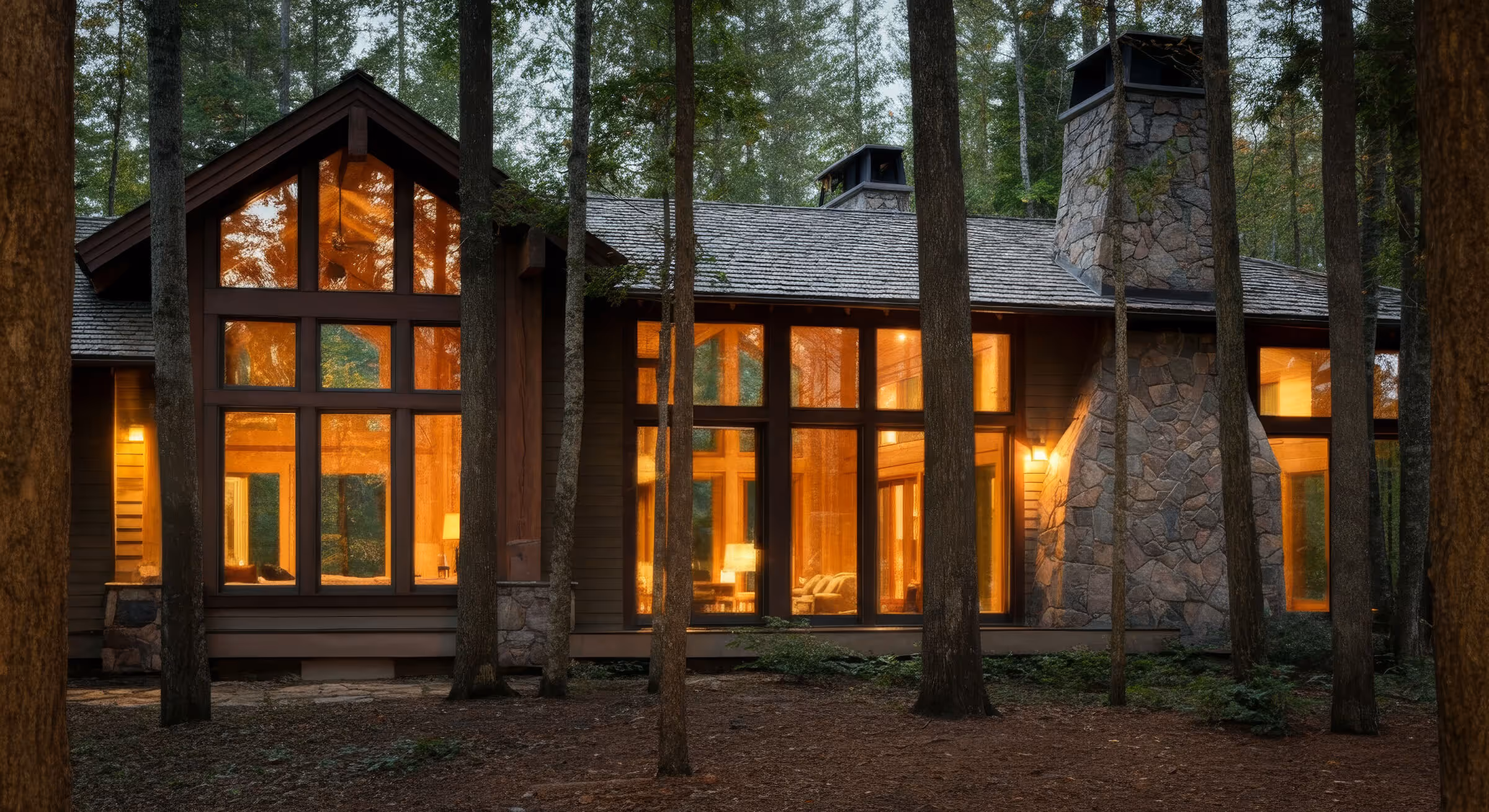 Modern cabin with large glowing windows and stone chimney surrounded by tall trees in a forest at dusk.