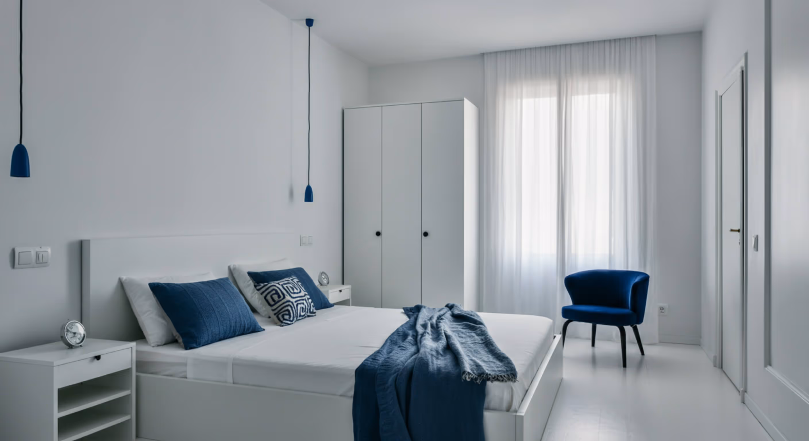 Minimalist bedroom with a white bed, blue pillows, blue throw blanket, two pendant lights, white wardrobe, and blue chair by a window with sheer white curtains.