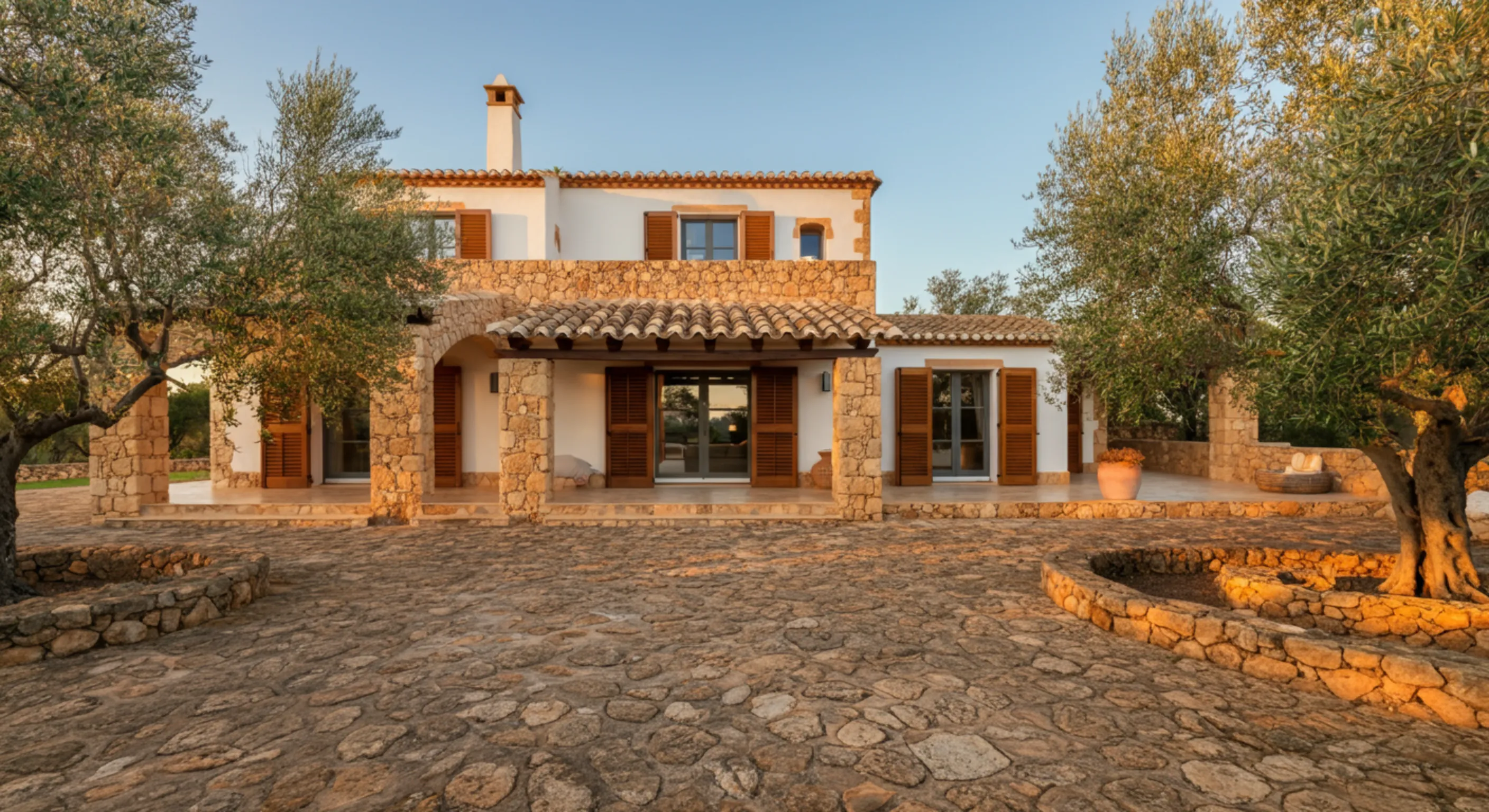 Stone and white stucco Mediterranean-style house with wooden shutters, terracotta roof tiles, surrounded by olive trees and stone patios.