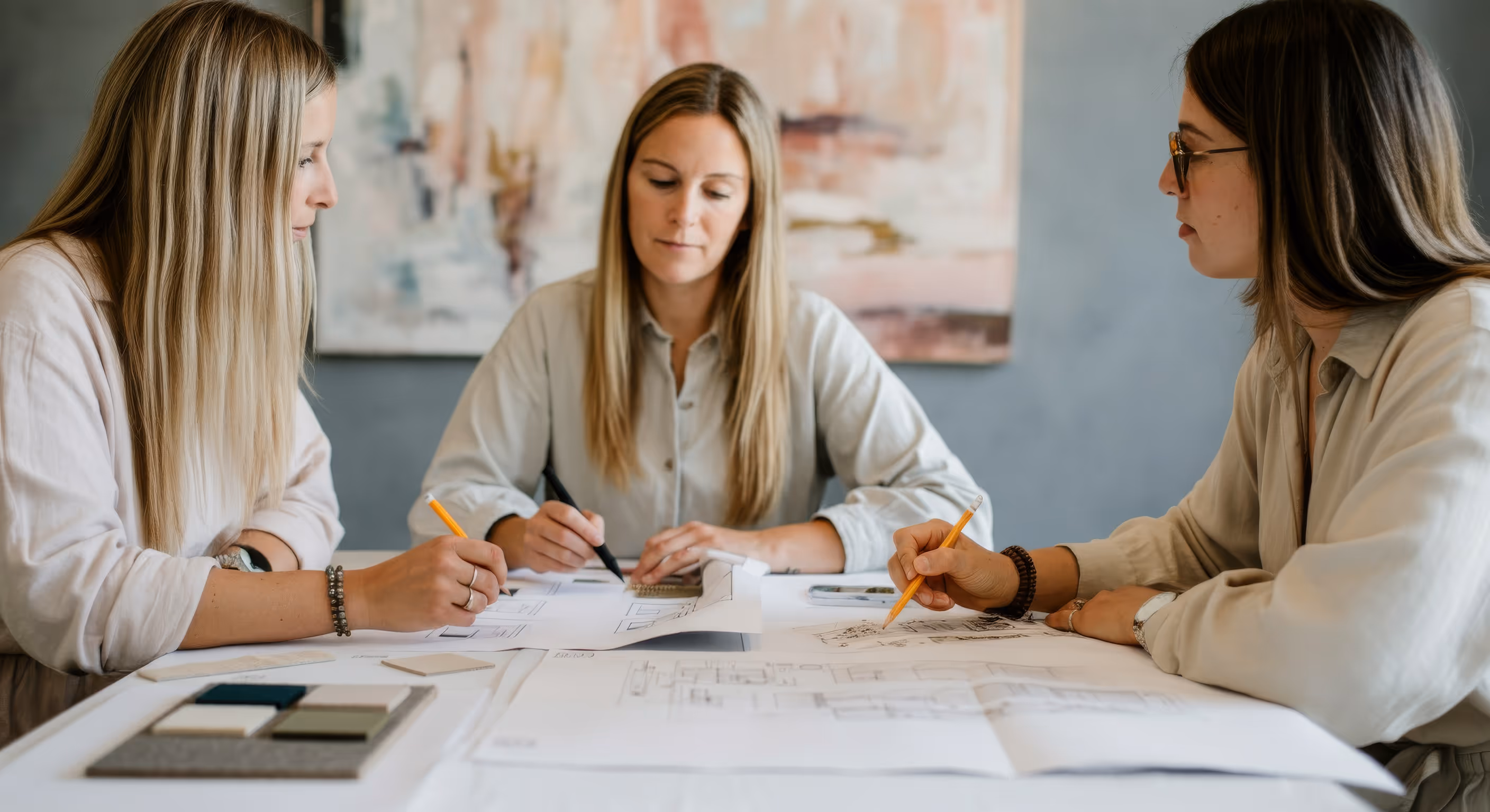 Three women sitting at a table reviewing architectural blueprints and design samples, each holding a pencil and engaged in discussion.