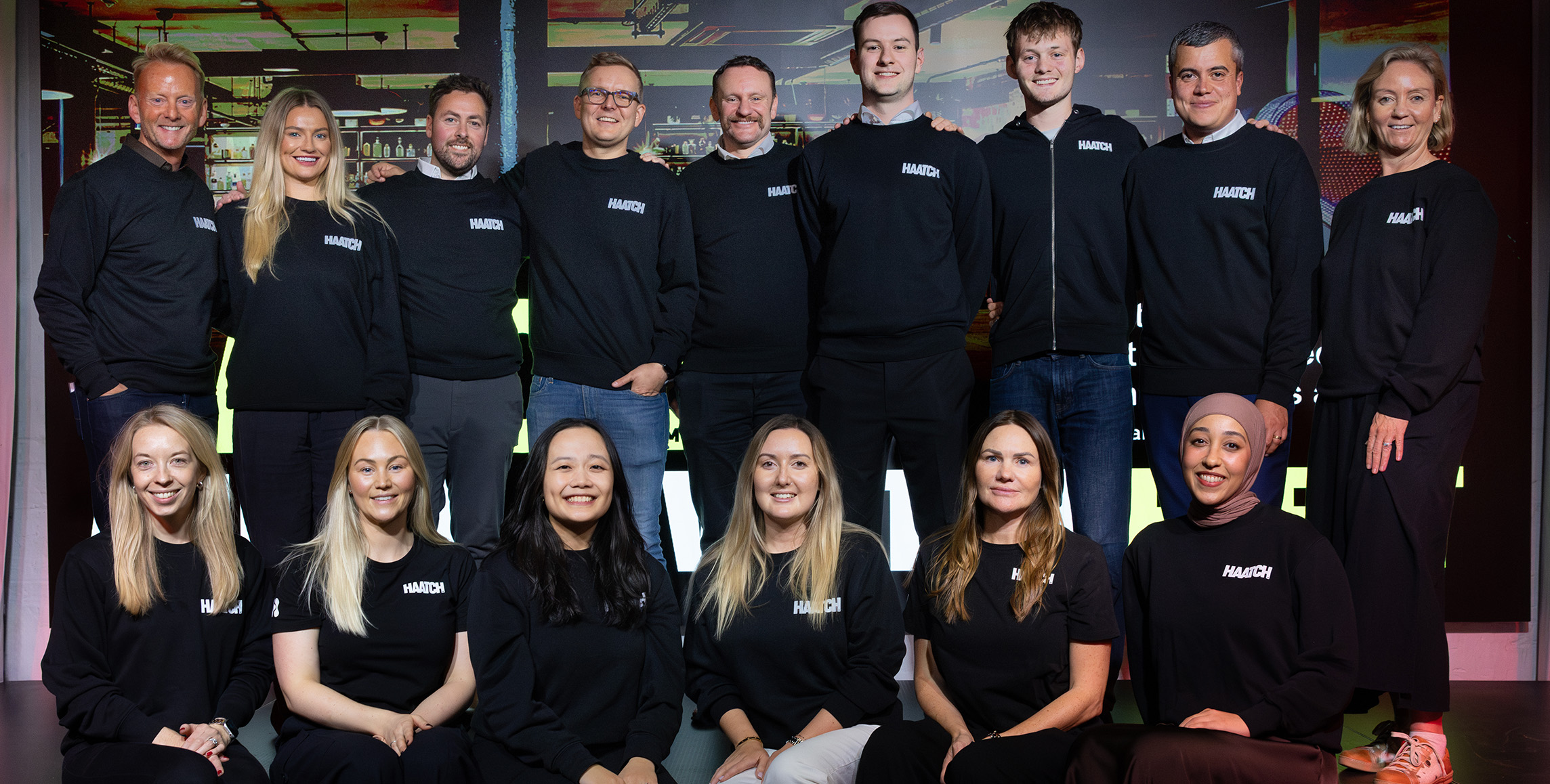 Group photo of sixteen smiling team members wearing black Haatch-branded sweatshirts and t-shirts in two rows.