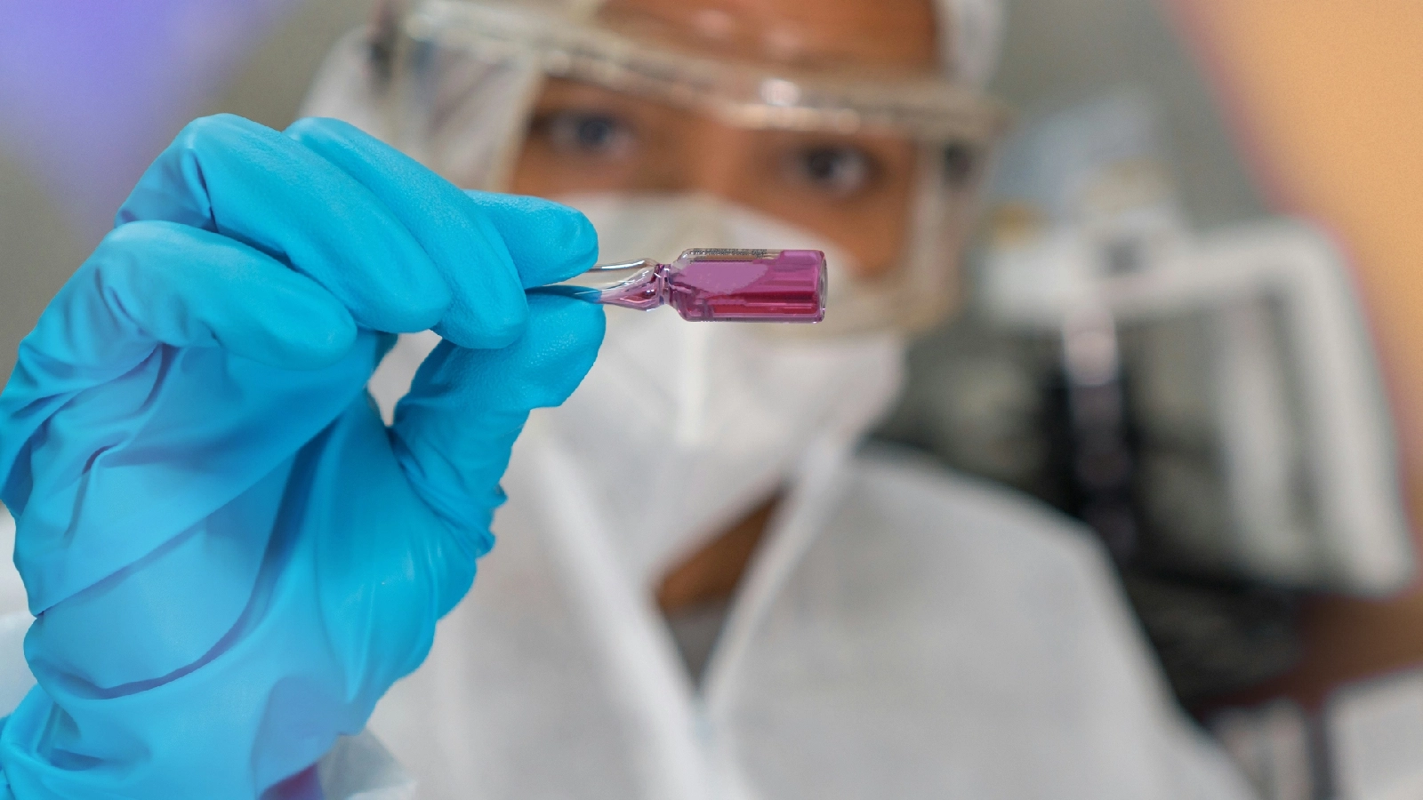 Scientist in protective gear holding a small vial of purple liquid with gloved hand