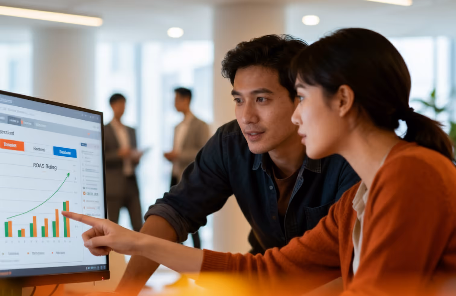 Two colleagues looking at a computer screen displaying a rising bar and line graph labeled 'ROAS Rating,' with one person pointing at the graph.