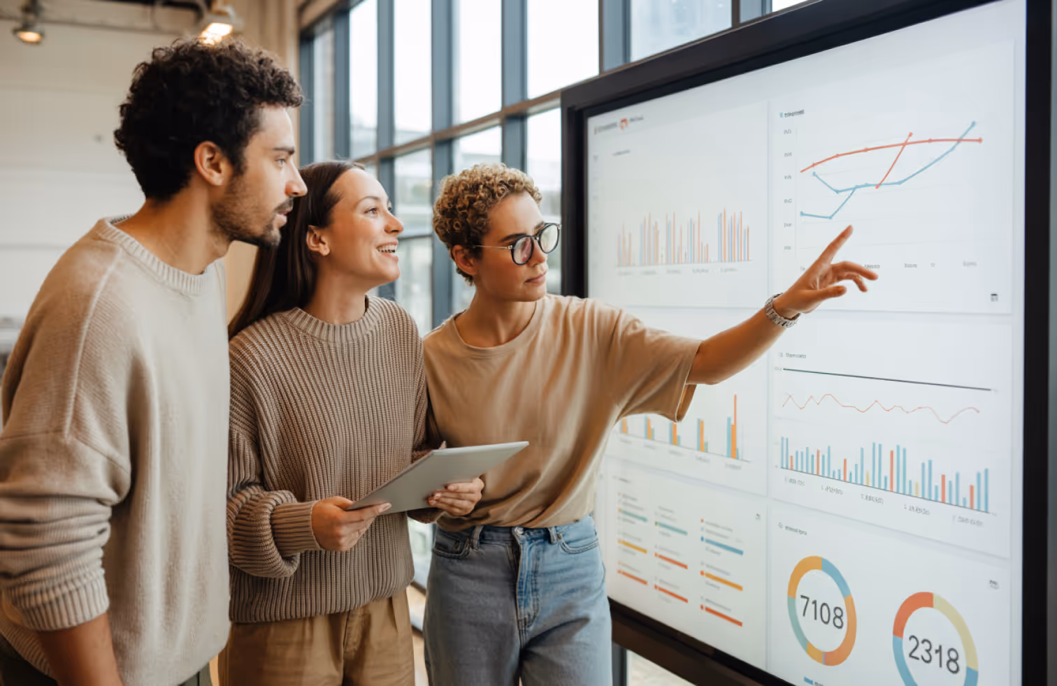 Three colleagues standing and discussing data charts on a large digital display in an office.