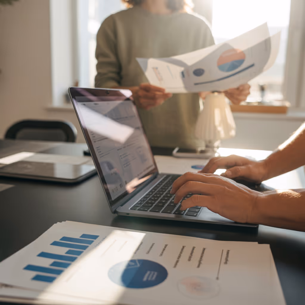 Person typing on a laptop with another person holding printed charts in the background, and documents with blue graphs on a table.