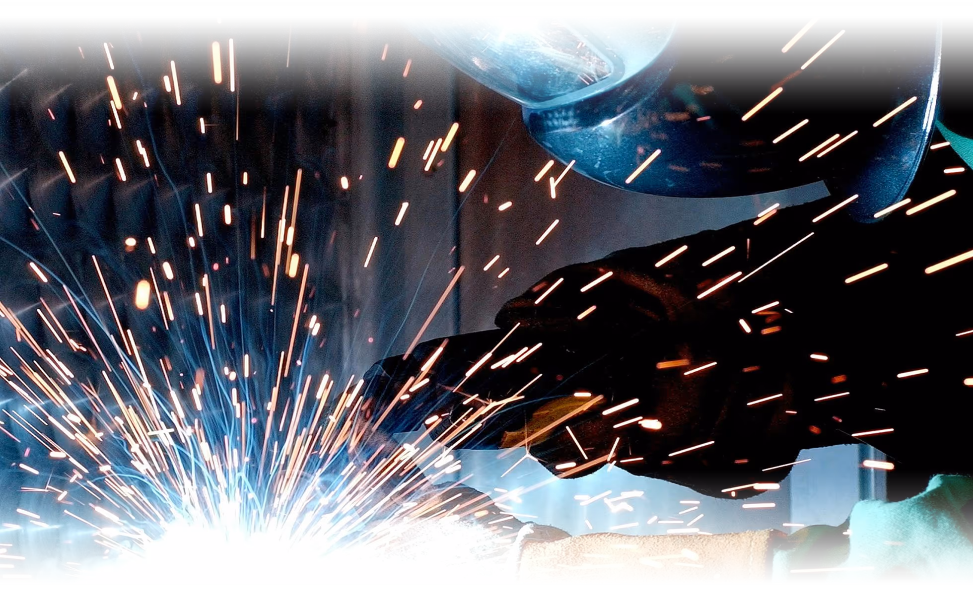 Welding in progress with a gloved hand guiding the torch, bright blue‑orange sparks flying against a dark workshop background, and a protective helmet partially visible.