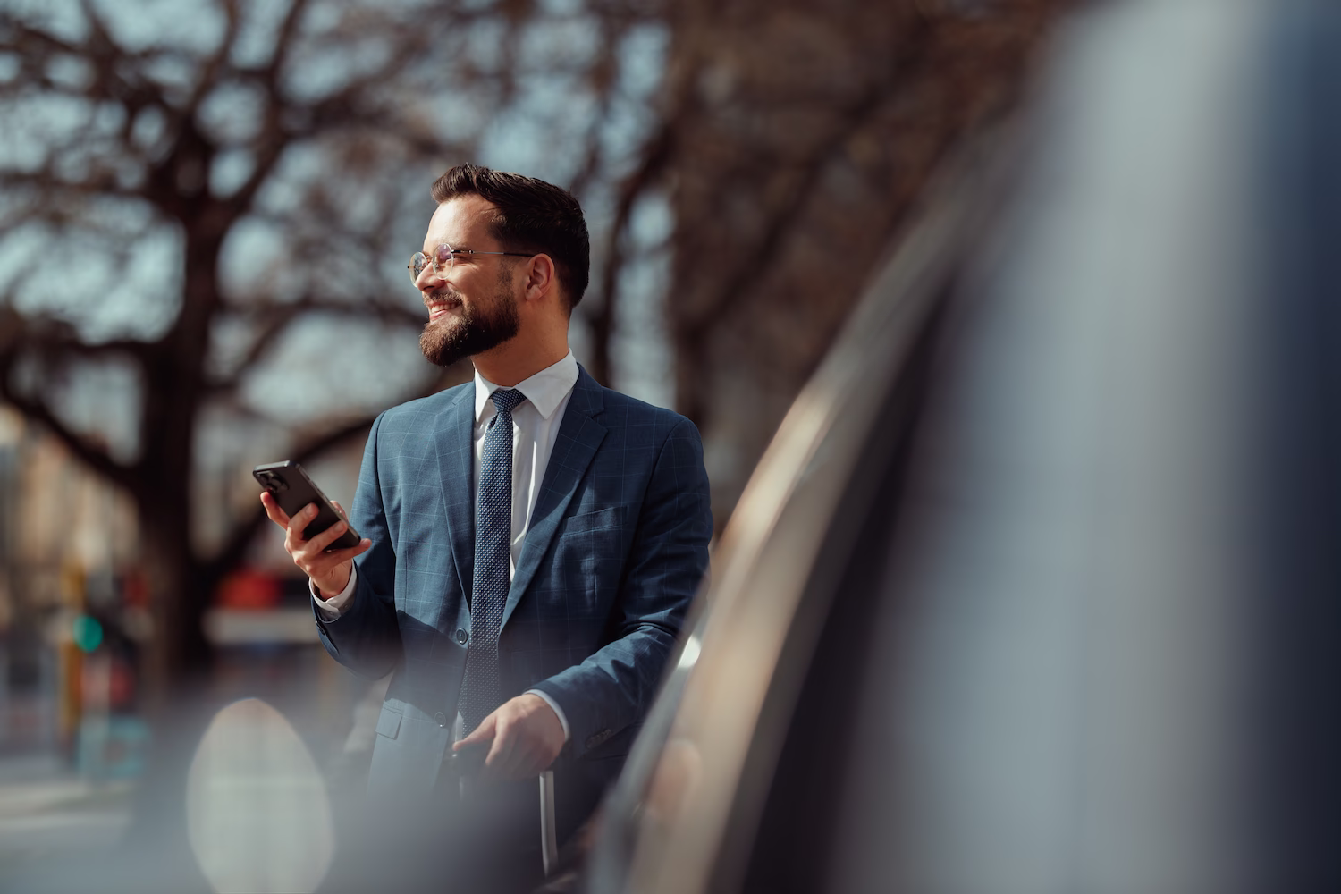 Homme d'affaires souriant en costume bleu avec téléphone et valise en plein air devant des arbres flous.