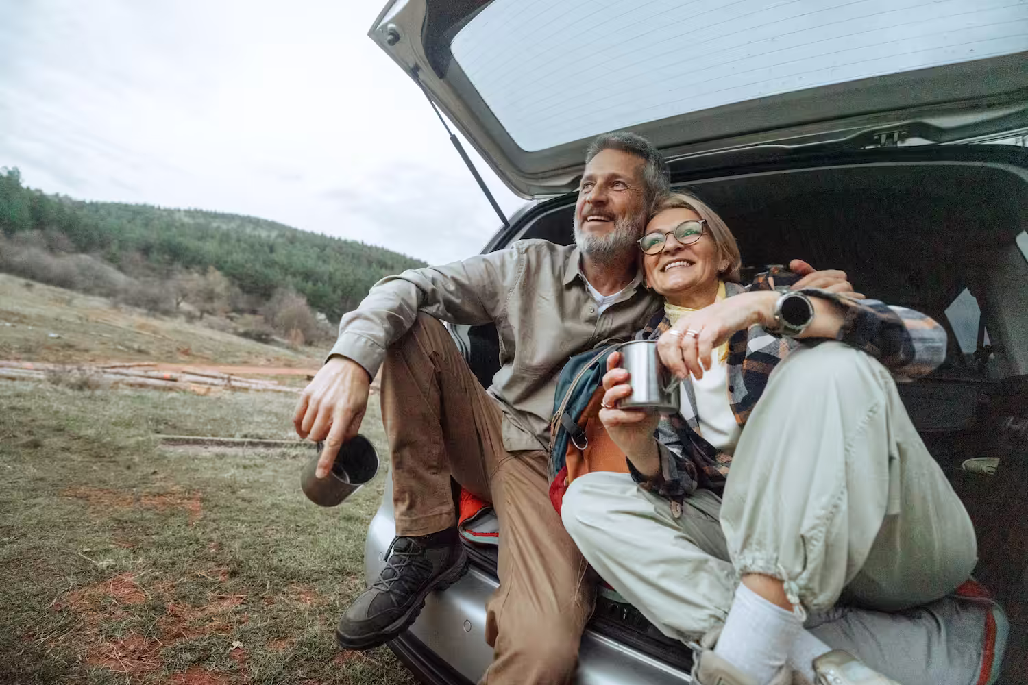 Un couple souriant assis à l'arrière d'une voiture avec des tasses en métal, entouré de nature.