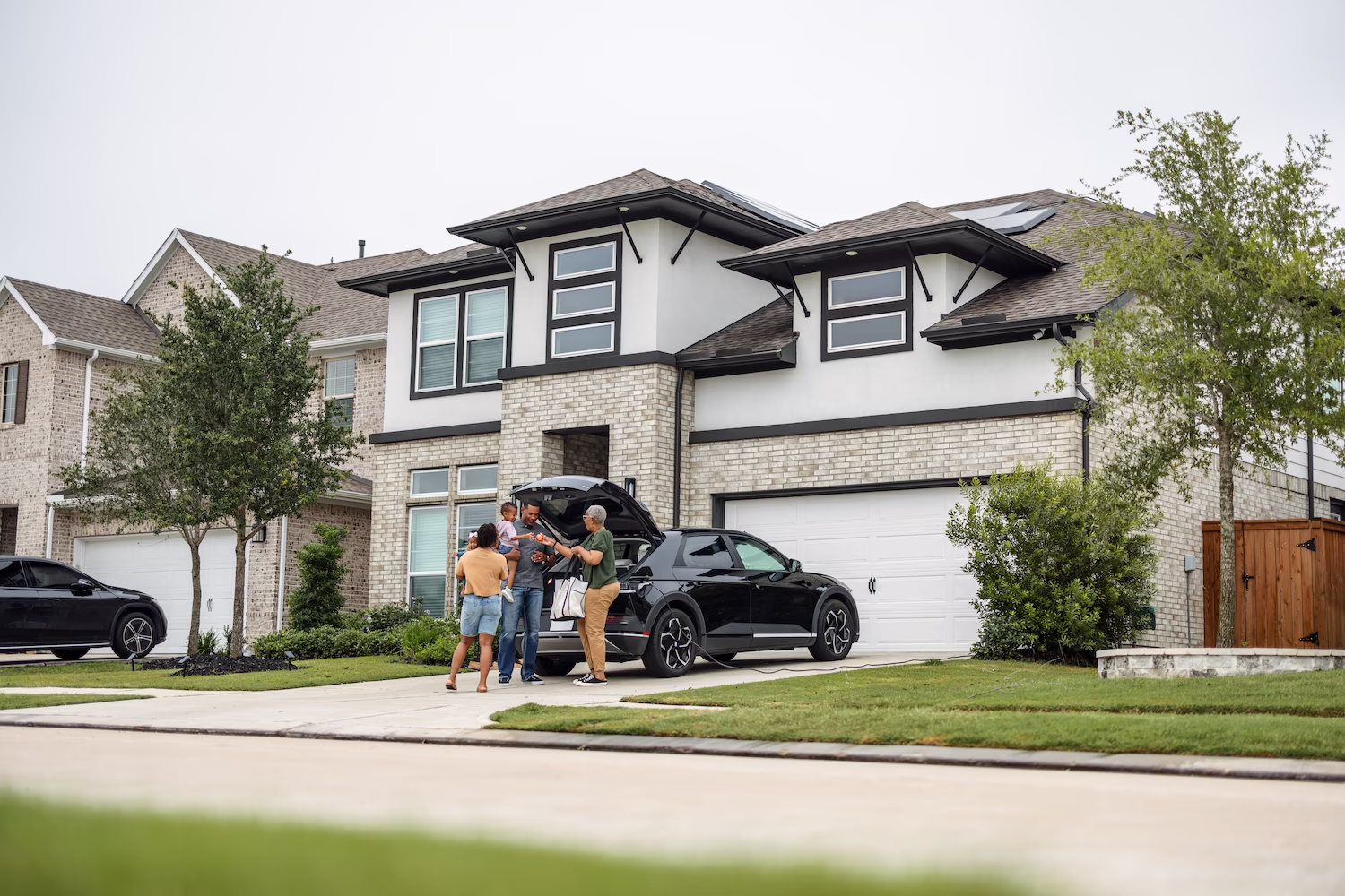 Une famille échangeant des objets près d'une voiture électrique noire en train d'être rechargée devant une maison moderne à deux étages.