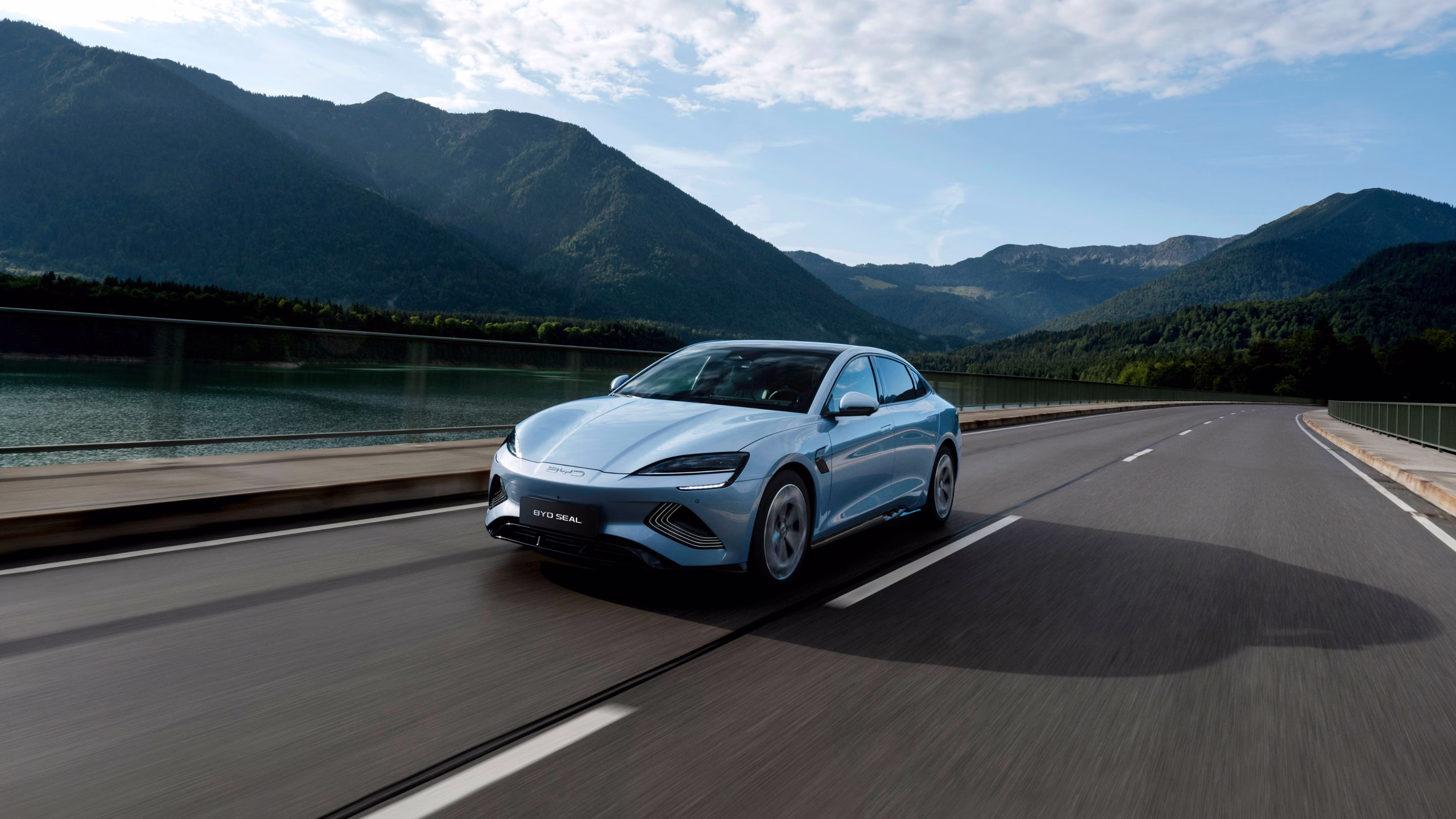 Voiture électrique BYD Seal bleu roulant sur une route près d'un lac avec des montagnes verdoyantes en arrière-plan.