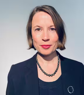 Porträt einer Frau mit kurzem braunem Haar, rotem Lippenstift und Perlenkette vor hellem Hintergrund.