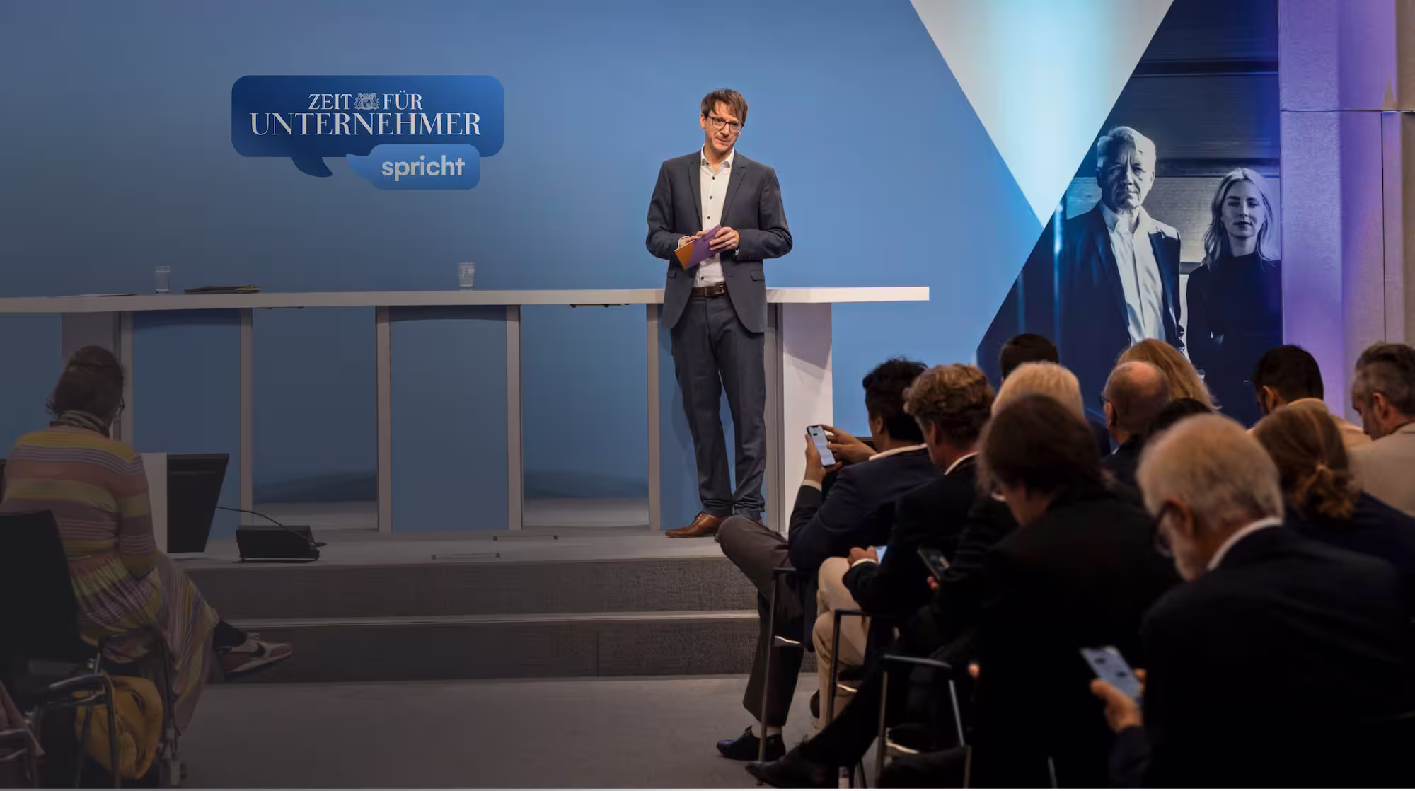 Ein Mann in Anzug steht auf einer Bühne vor einem blauen Hintergrund mit der Aufschrift 'Zeit für Unternehmer spricht', während das Publikum im Vordergrund sitzt.
