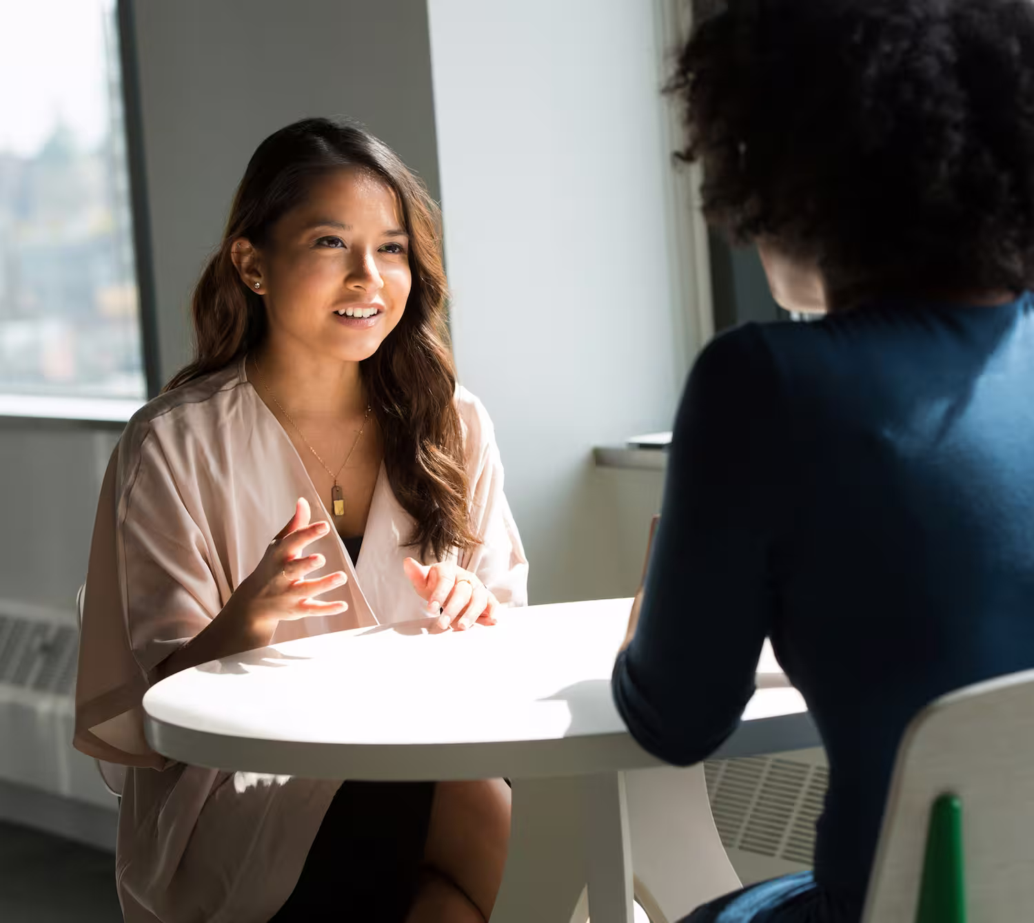 Zwei Frauen im Gespräch an einem runden weißen Tisch in einem hellen Raum mit Fenster.