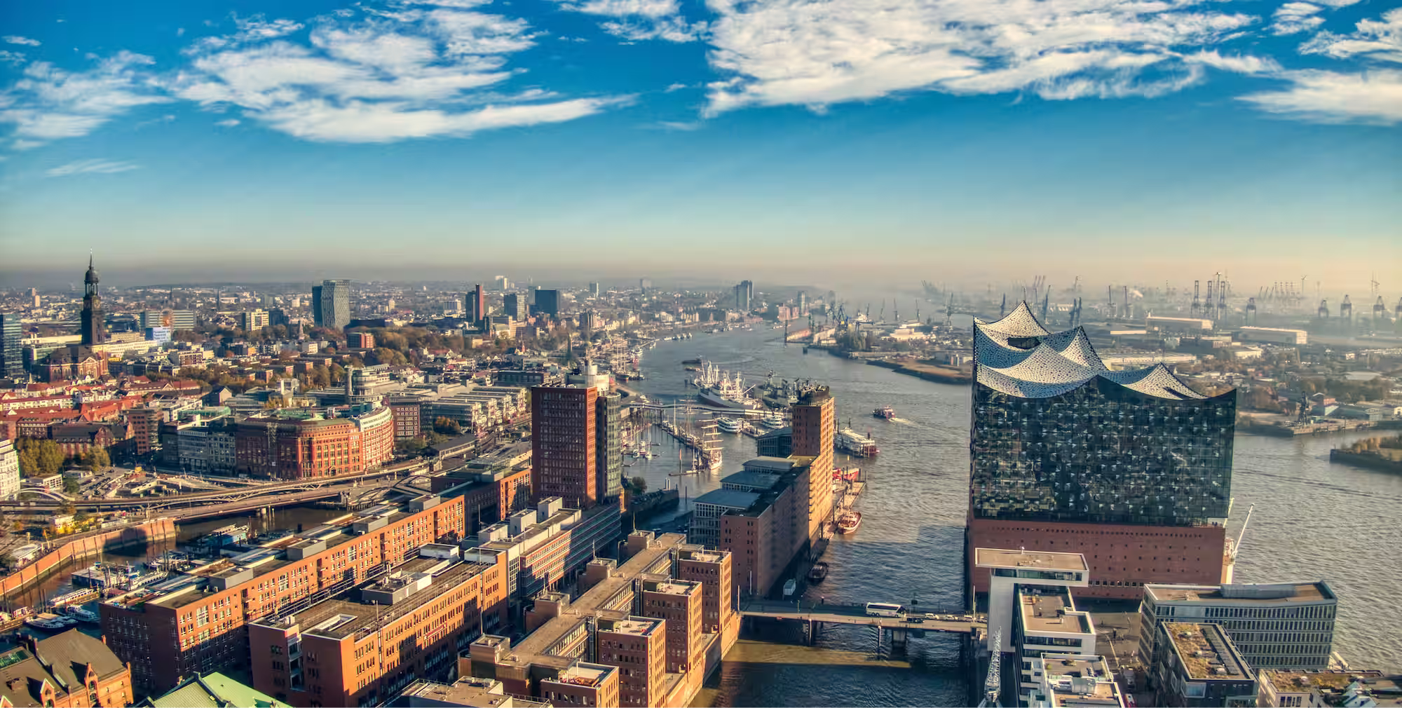 Luftaufnahme des Hamburger Hafens mit der Elbphilharmonie rechts und zahlreichen Gebäuden entlang des Flusses unter blauem Himmel.
