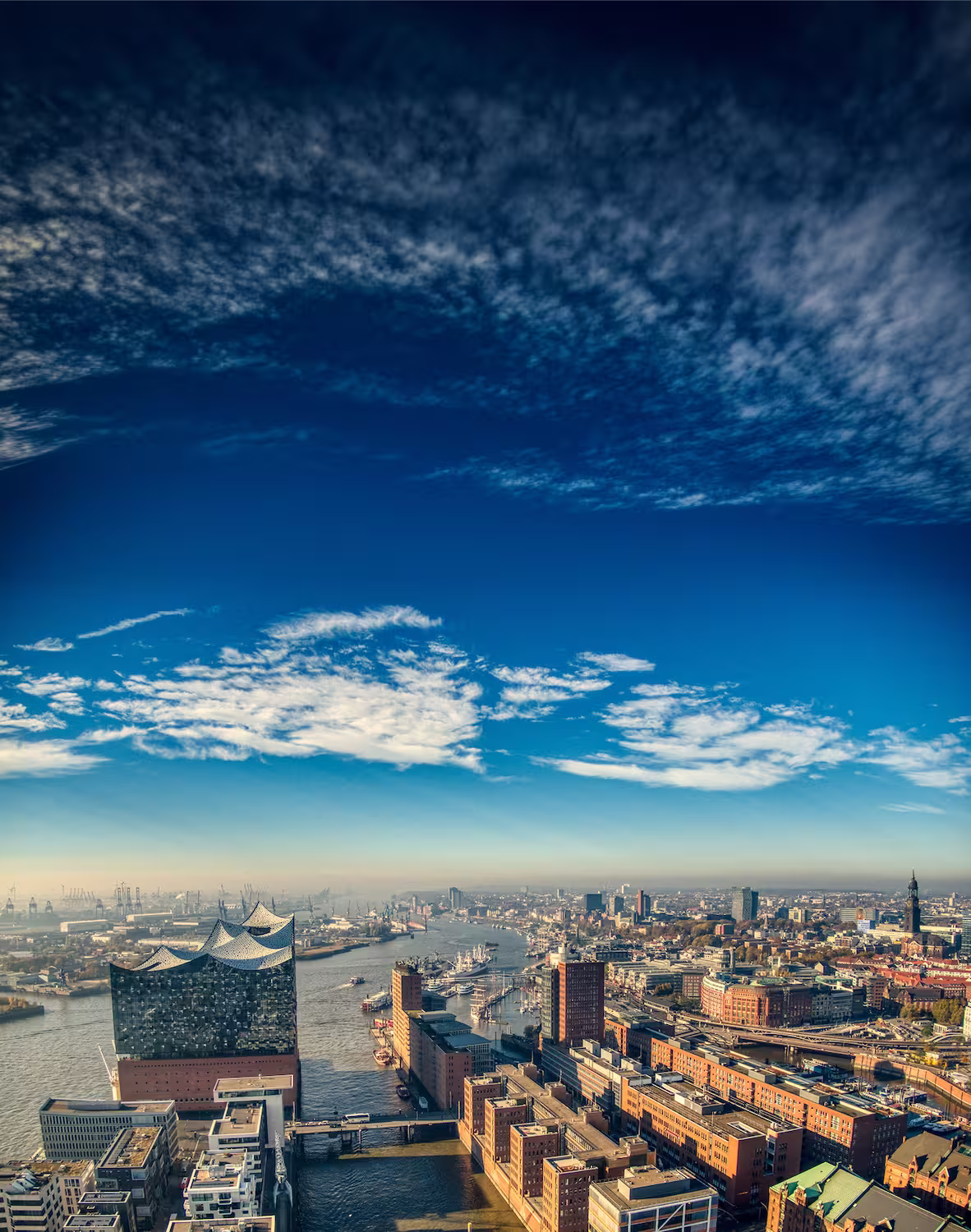 Luftaufnahme des Hamburger Hafens mit der Elbphilharmonie und der Elbe unter einem bewölkten blauen Himmel.