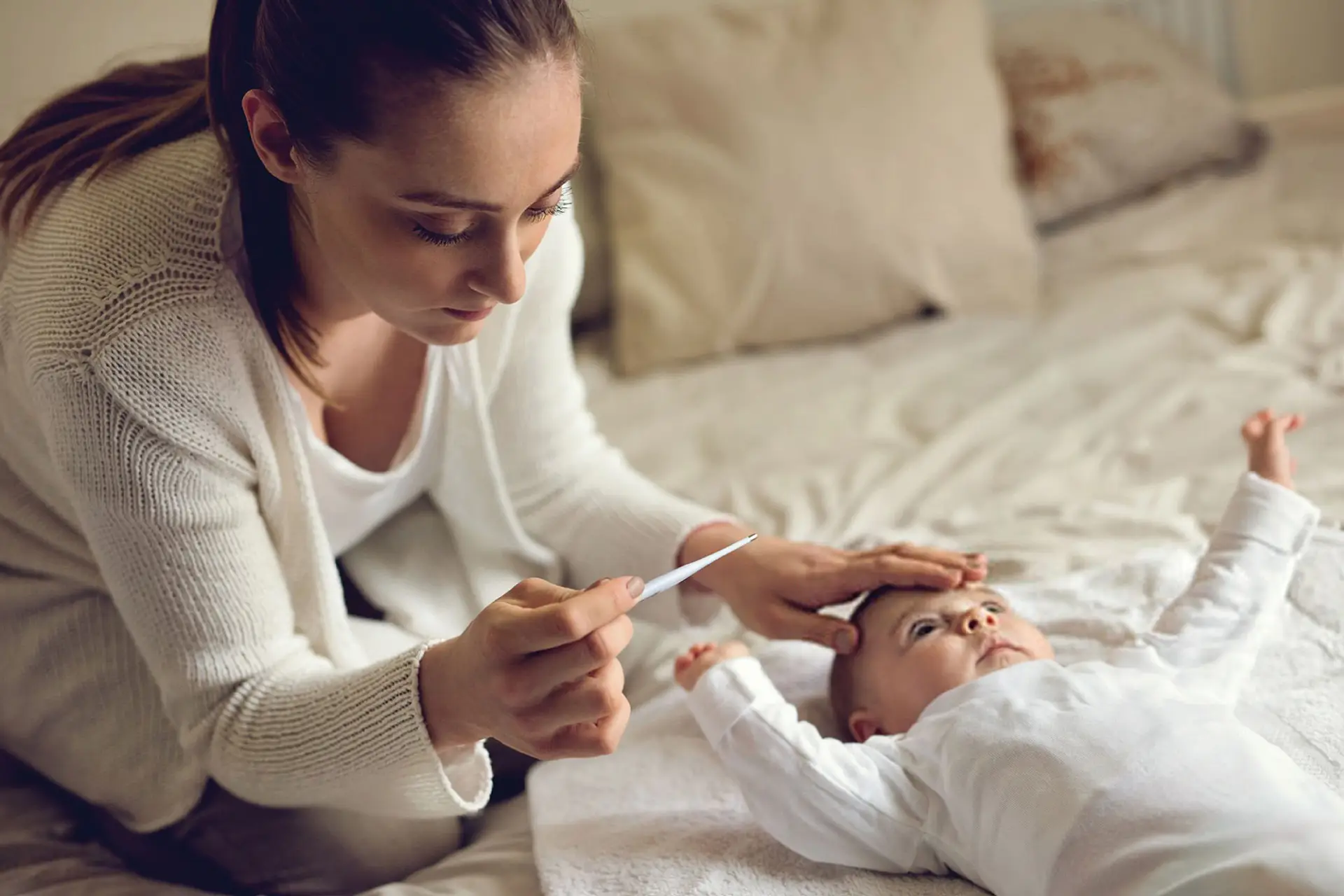 Parent checking a baby’s temperature with a thermometer while the baby lies on a bed.