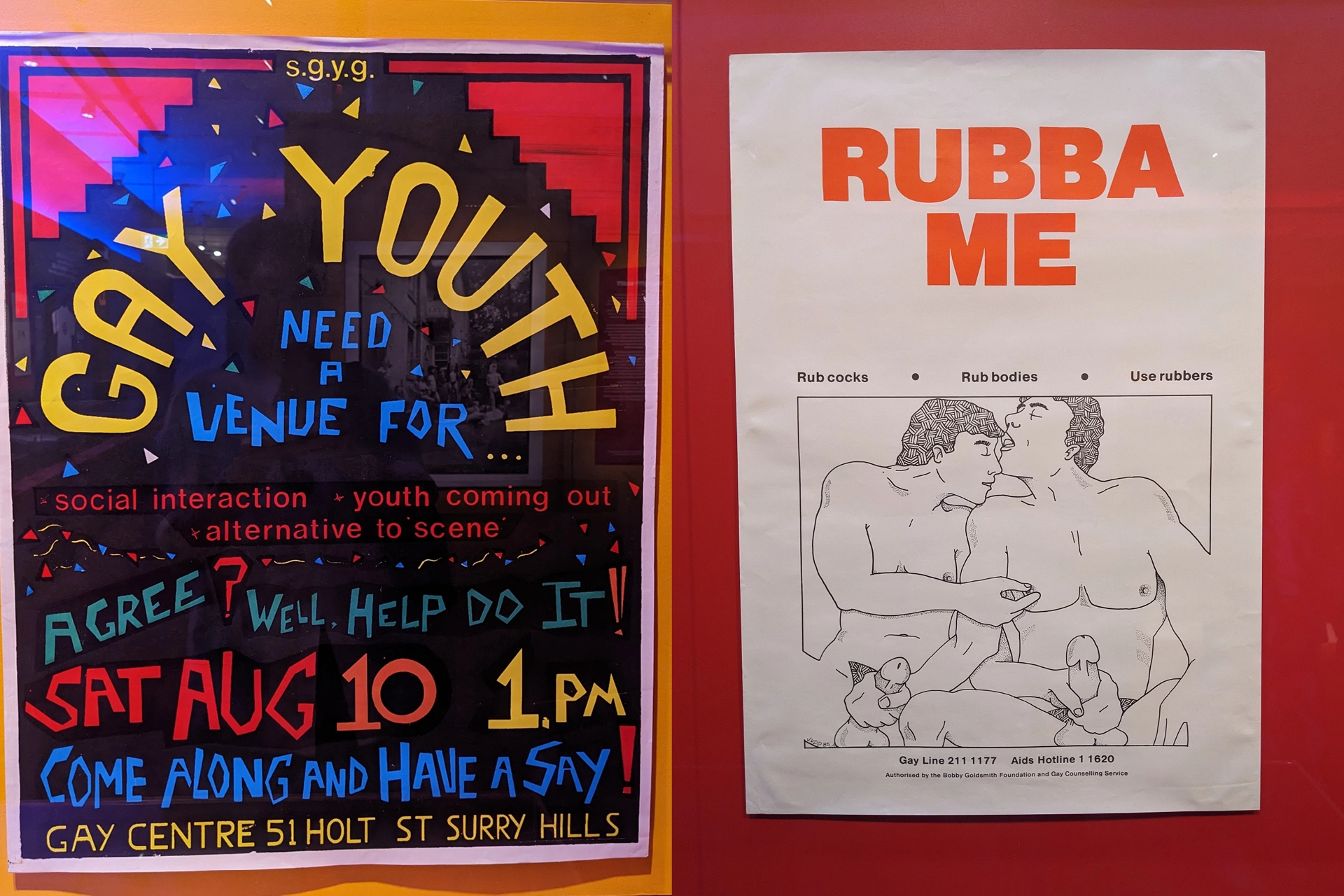 Historic queer posters displayed in the exhibition, including a ‘Gay Youth’ community event poster and a ‘Rubba Me’ illustrated design.