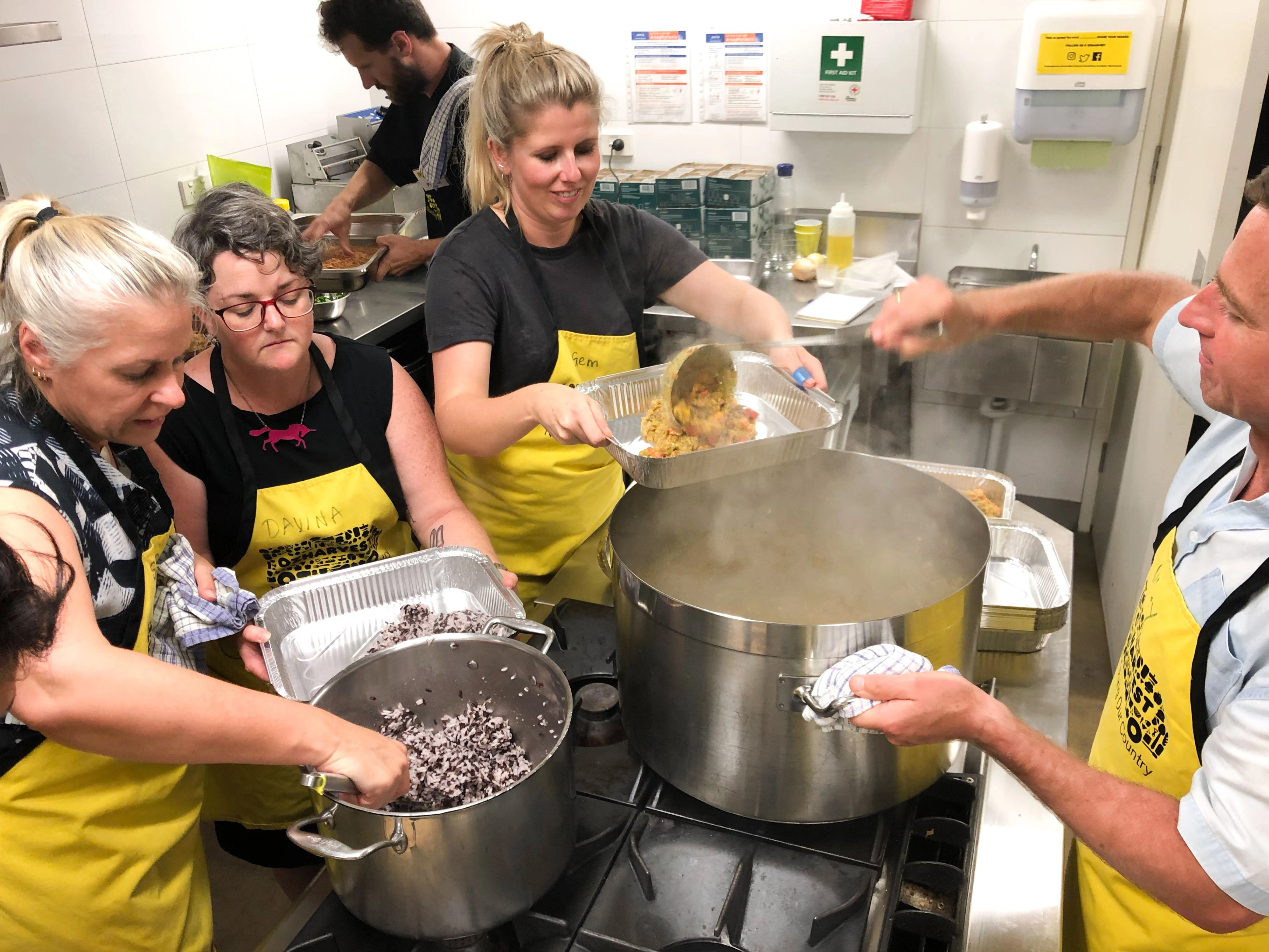Folk team members in yellow OzHarvest aprons preparing and portioning food from large pots in a commercial kitchen.