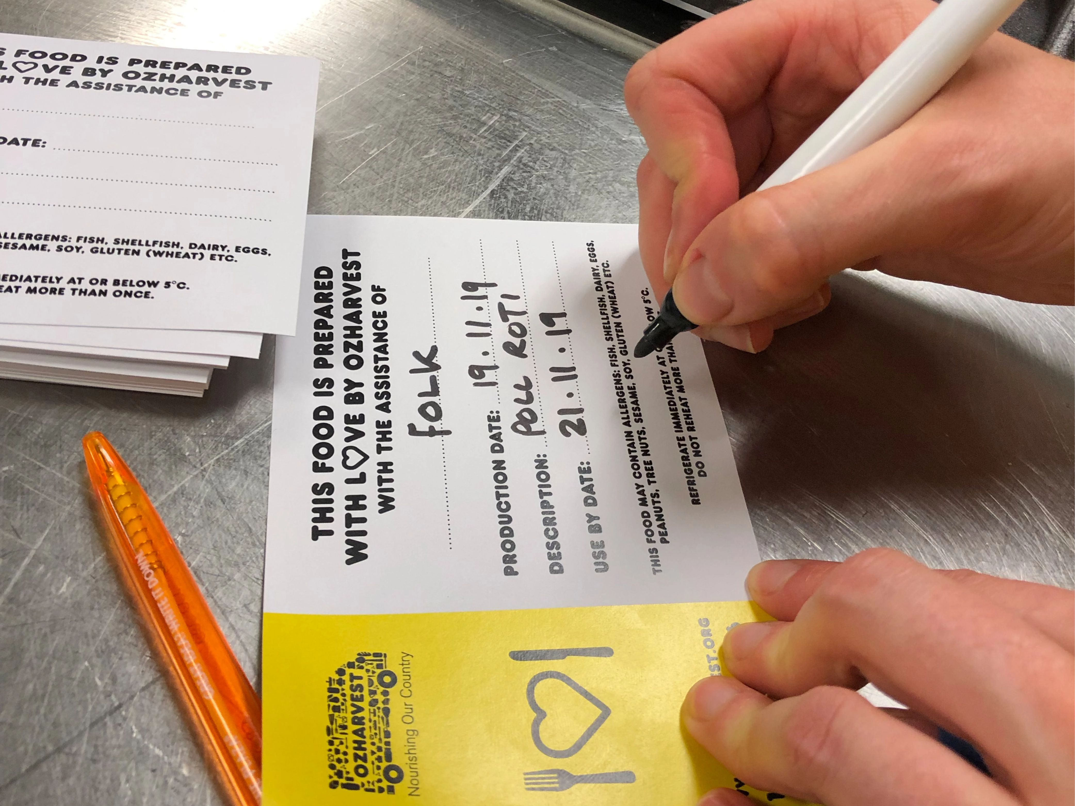 Close-up of a participant writing a meal label that reads ‘This food is prepared with love by OzHarvest with the assistance of Folk.’