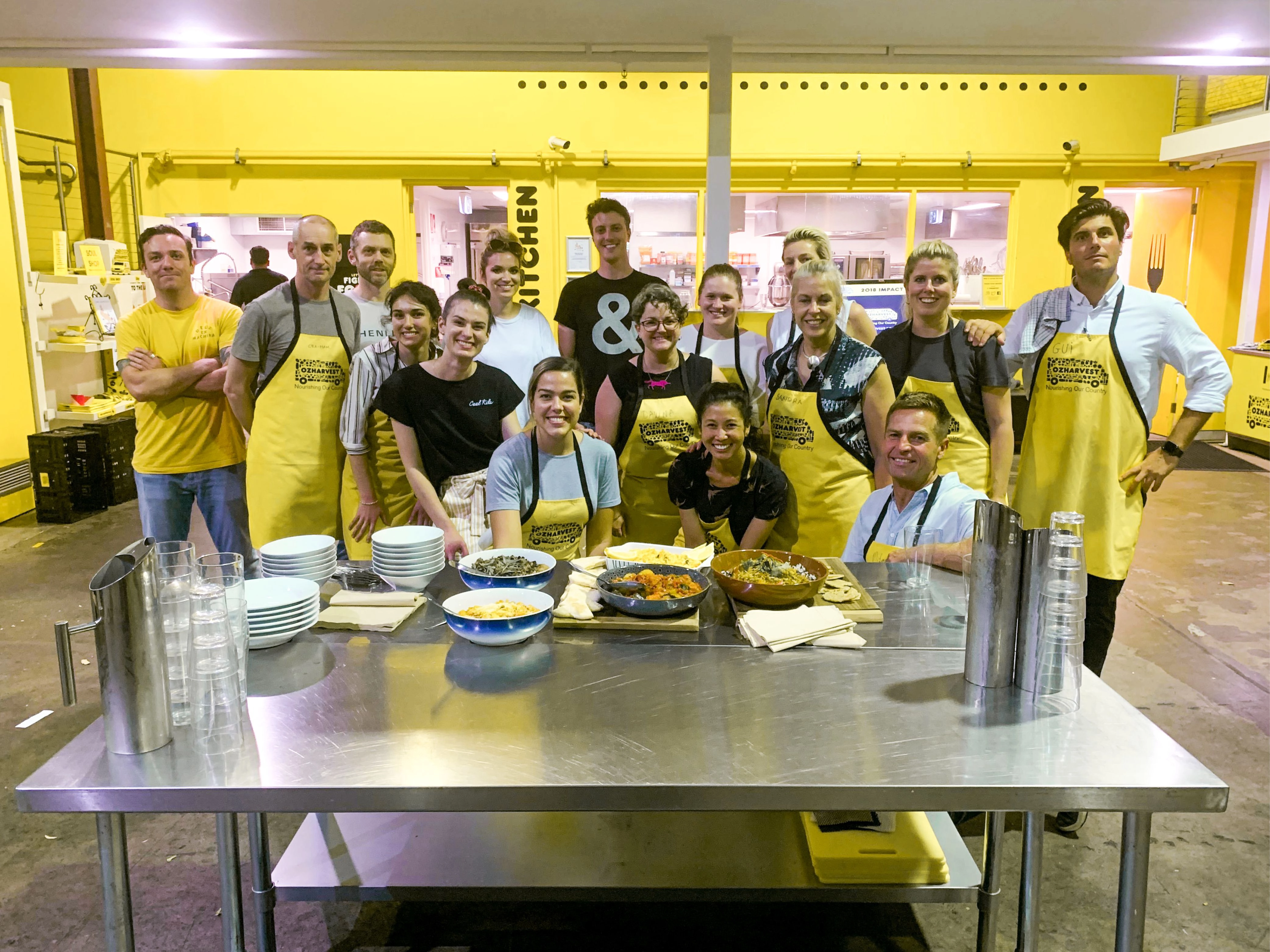 The Folk team wearing yellow OzHarvest aprons gathered around a table of completed meals at the end of the Cooking for a Cause event.