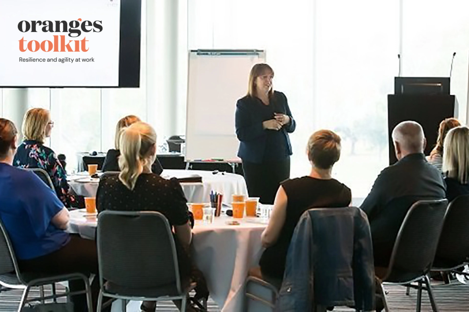A facilitator from the Oranges Toolkit delivers a workplace resilience workshop to a group of employees seated around tables.