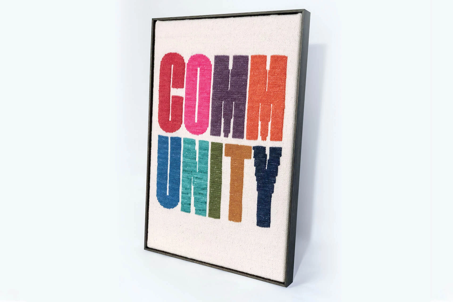 Framed woven artwork reading ‘COMMUNITY’ in bold multicoloured lettering.