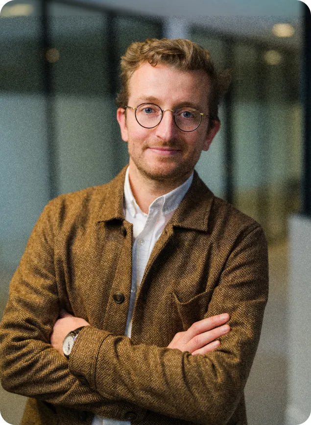 DAF avec lunettes, veste marron et chemise blanche, bras croisés, posant dans un espace de bureau moderne.