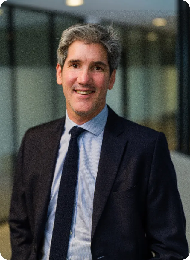 DAF souriant portant un costume bleu foncé avec une chemise à rayures et une cravate noire, debout devant une façade vitrée floue.