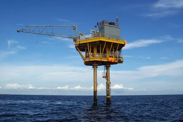 Small offshore oil platform with yellow support legs in calm ocean under blue sky.