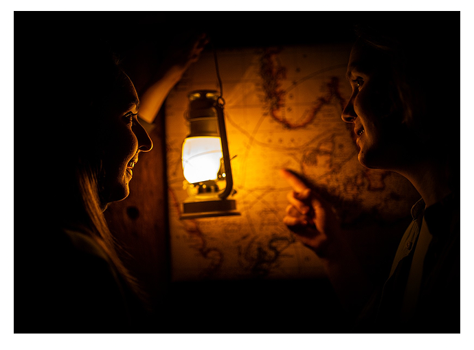 Two people illuminated by a lantern, smiling and pointing at a map on a wall in dim light.