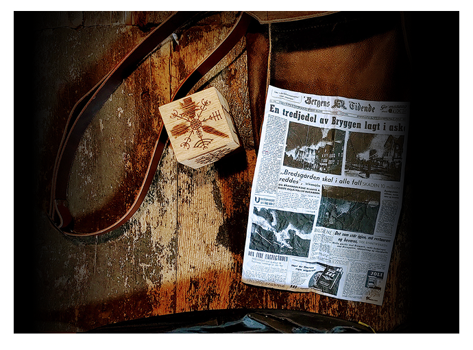 Old newspaper and a wooden block with carved symbols on a rustic wooden surface with a leather strap beside them.