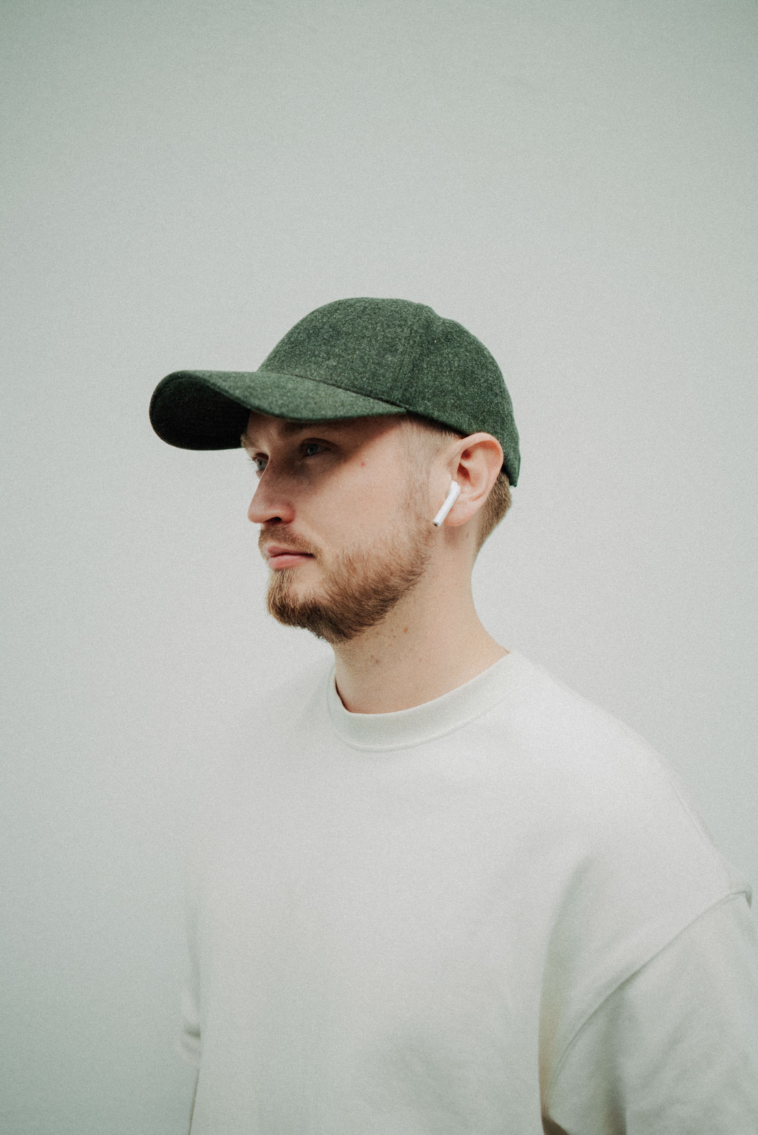 Young man with a beard wearing a green cap, white sweatshirt, and white wireless earbuds, looking to the left.