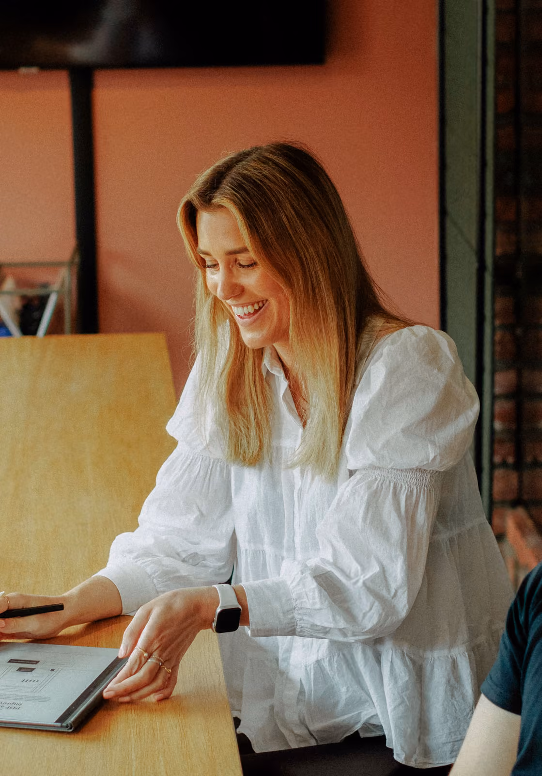Smiling woman with blonde hair in a white blouse looking at a tablet on a wooden table.