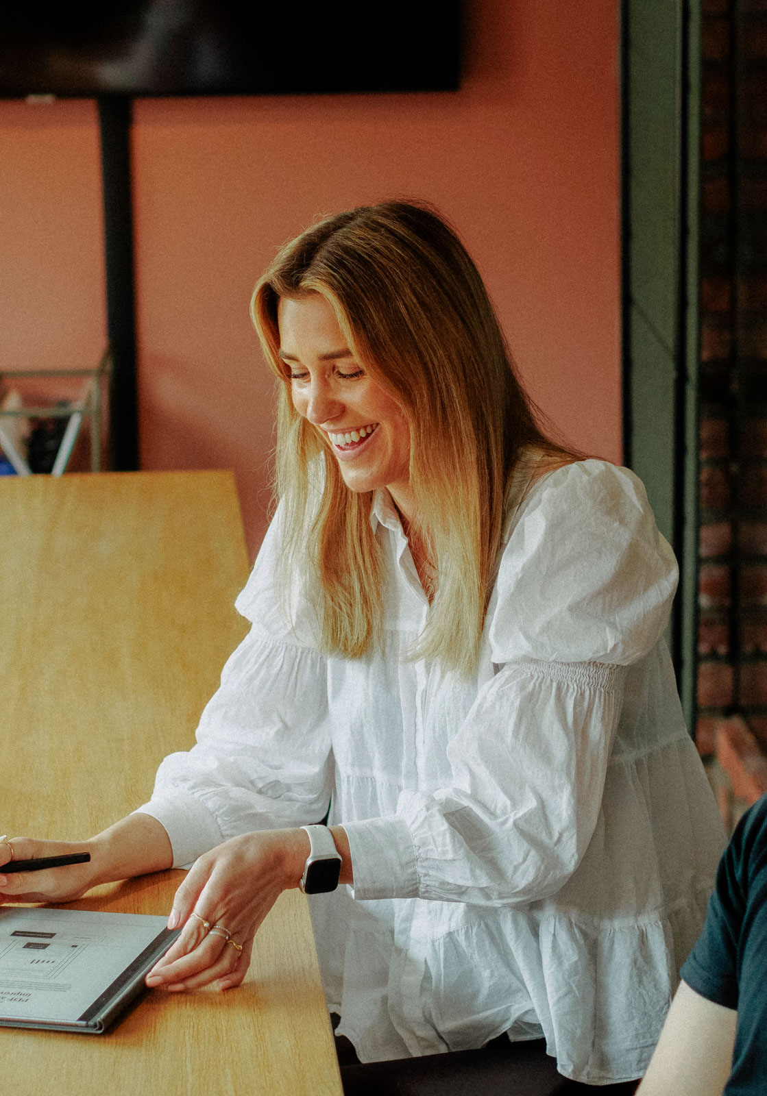 Smiling woman with blonde hair in a white blouse looking at a tablet on a wooden table.