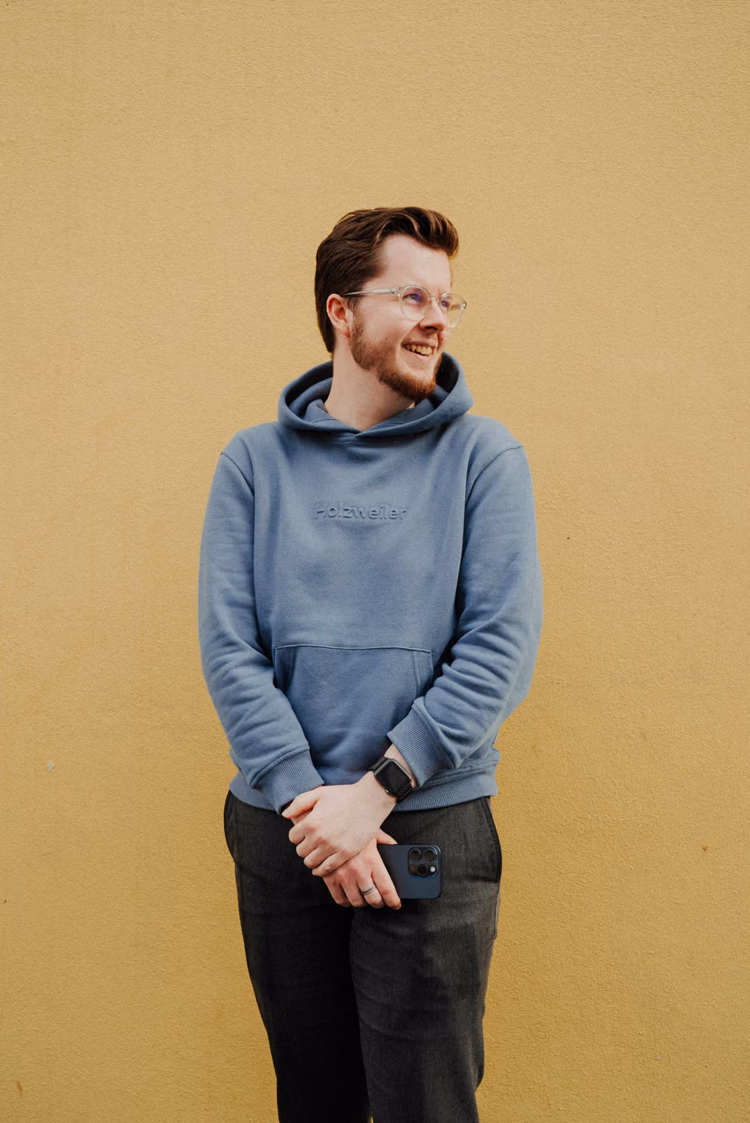 Smiling man with glasses wearing a blue hoodie holding a smartphone in front of a yellow wall.