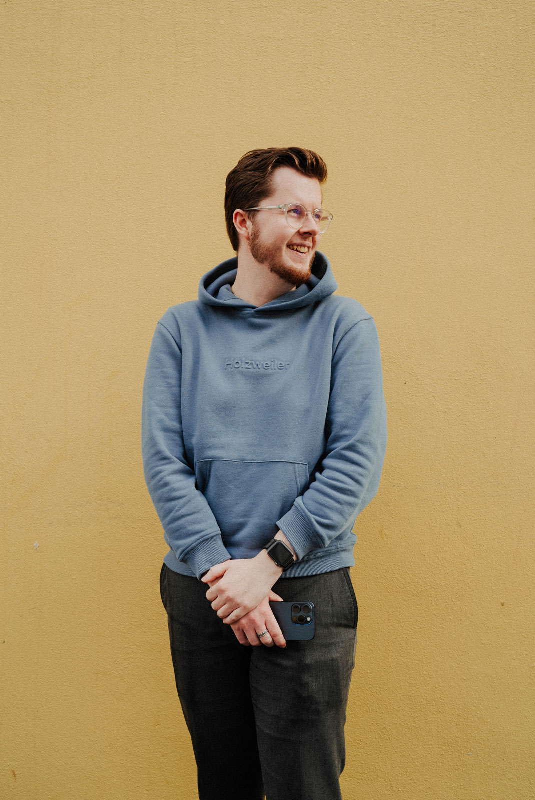 Smiling man with glasses wearing a blue hoodie holding a smartphone in front of a yellow wall.