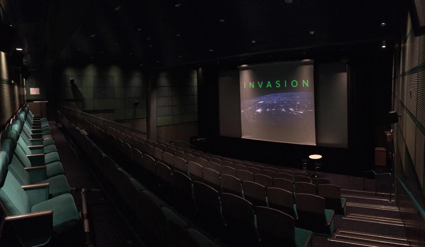 Empty cinema room with rows of seats facing a screen displaying the word 'INVASION' over an image of Earth at night.