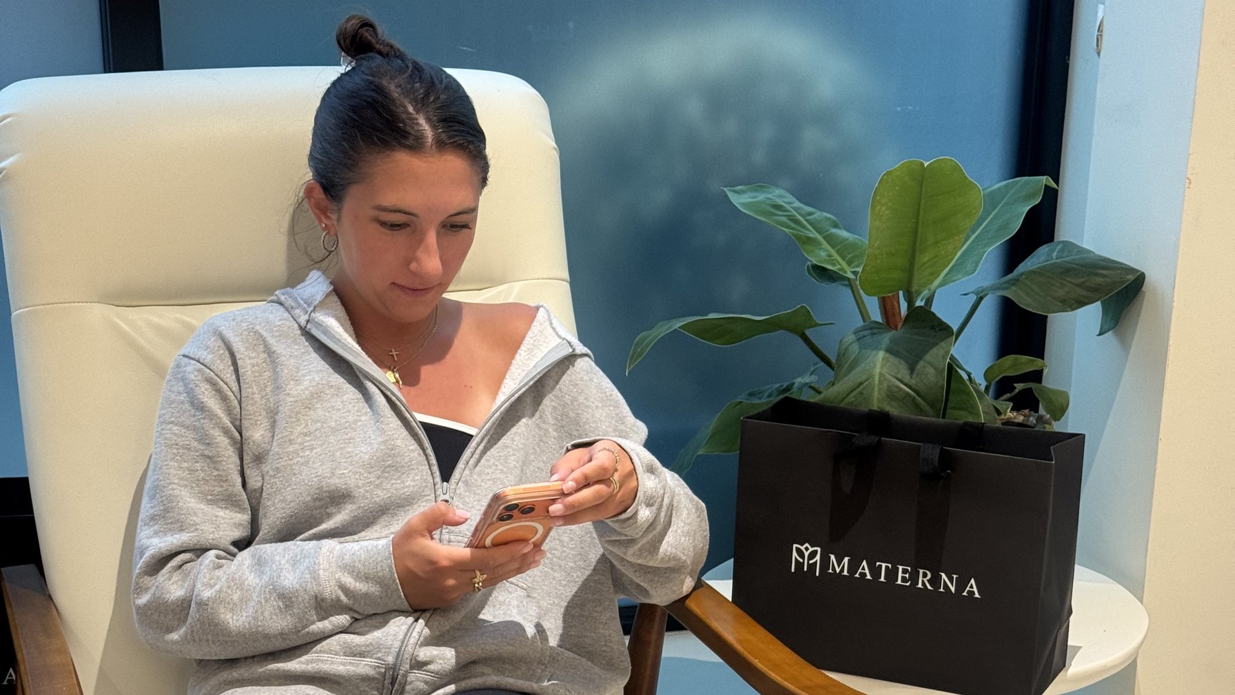 A woman sits in a cream armchair in the Materna waiting area in the West Village, looking at her phone, with a Materna tote bag on the side table beside her.