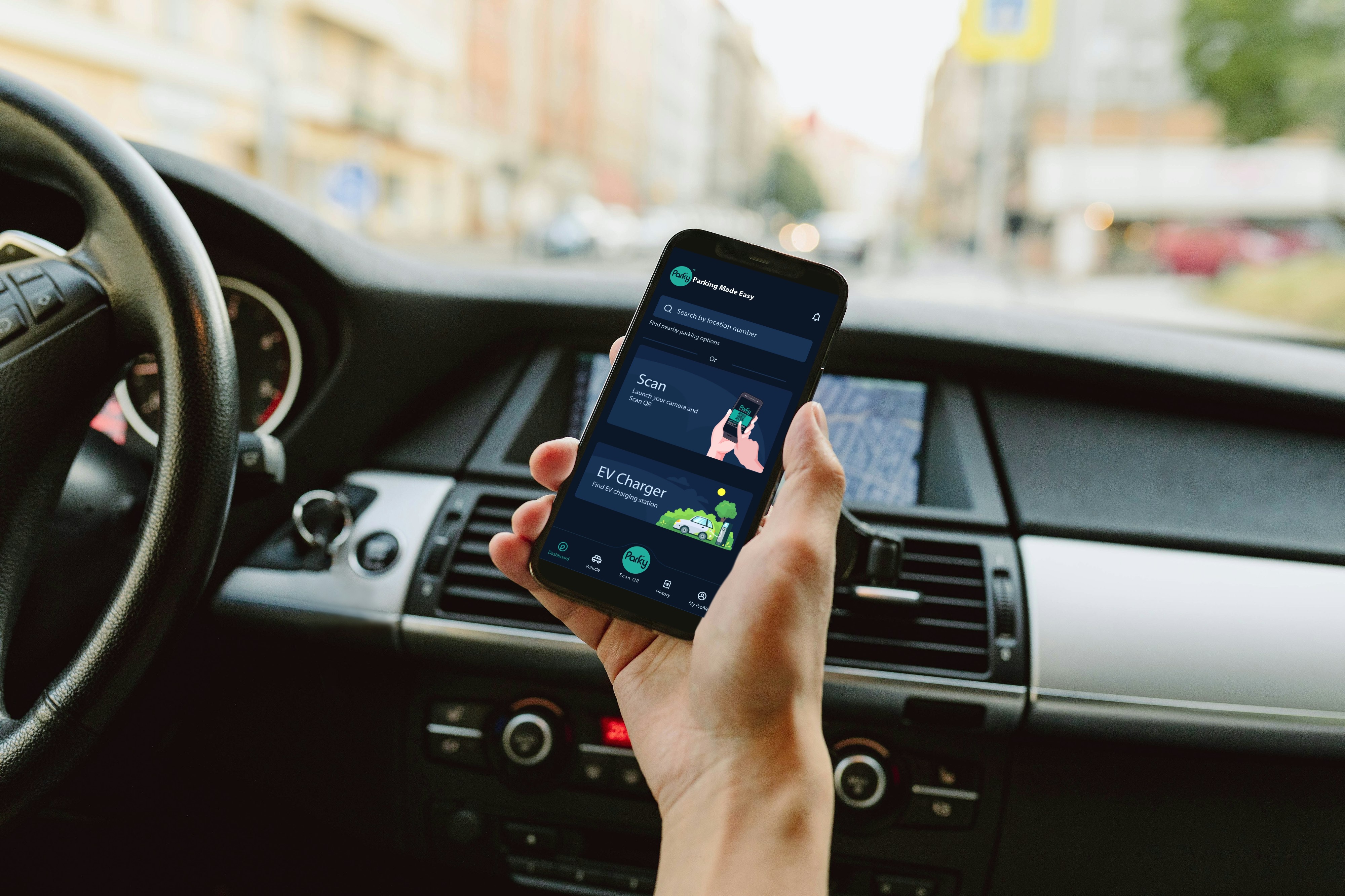Person holding a smartphone inside a car displaying a parking app interface with options to search, scan, and find EV chargers.