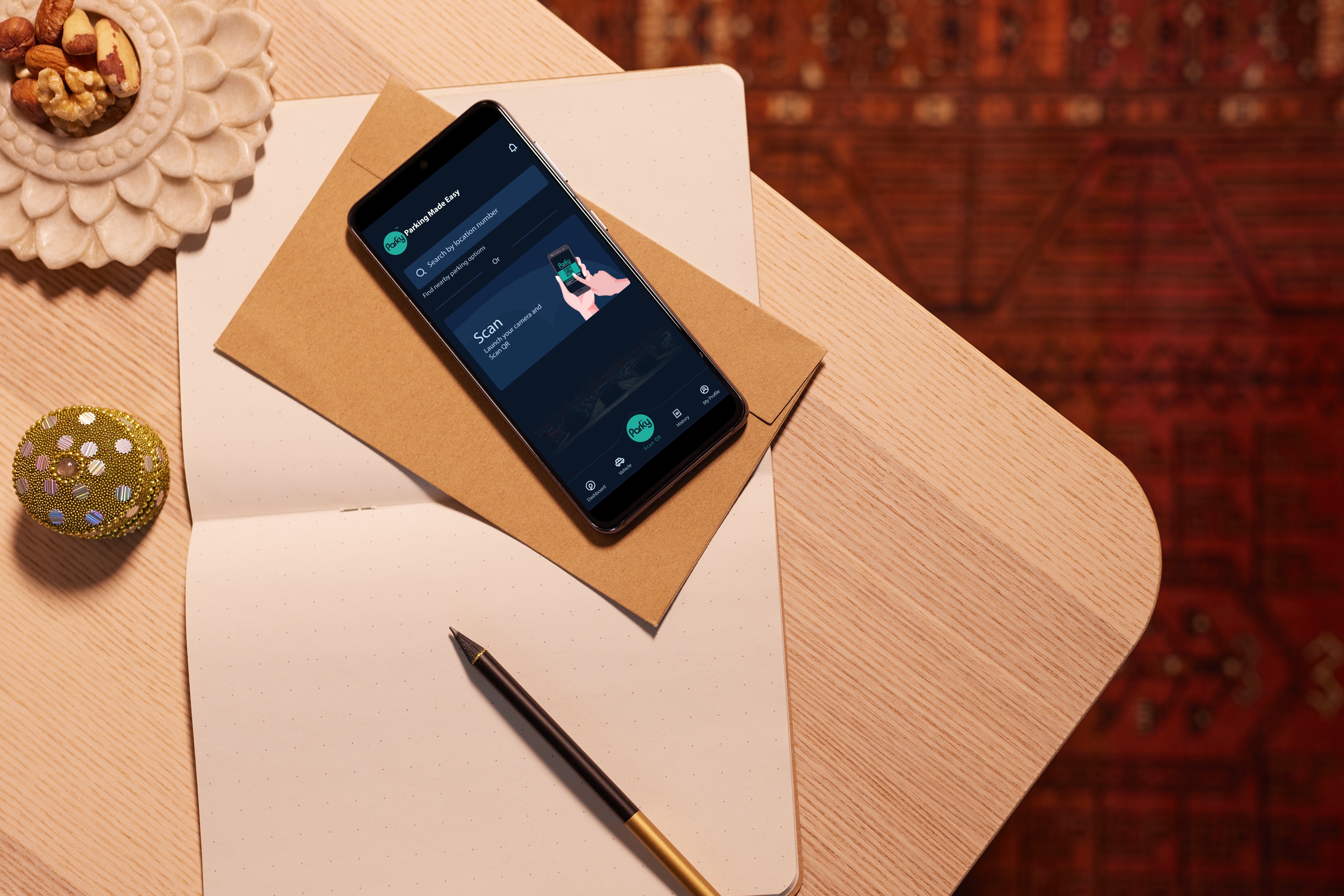 Smartphone on a brown envelope displaying a parking app screen, resting on an open dotted notebook with a pen on a wooden table, next to a bowl of mixed nuts and a decorative golden item.