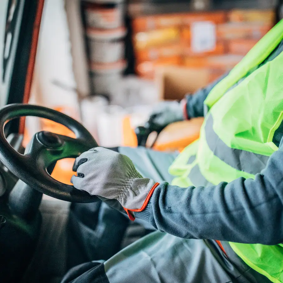 Worker in high-visibility vest and gloves operating a forklift steering wheel inside a warehouse.