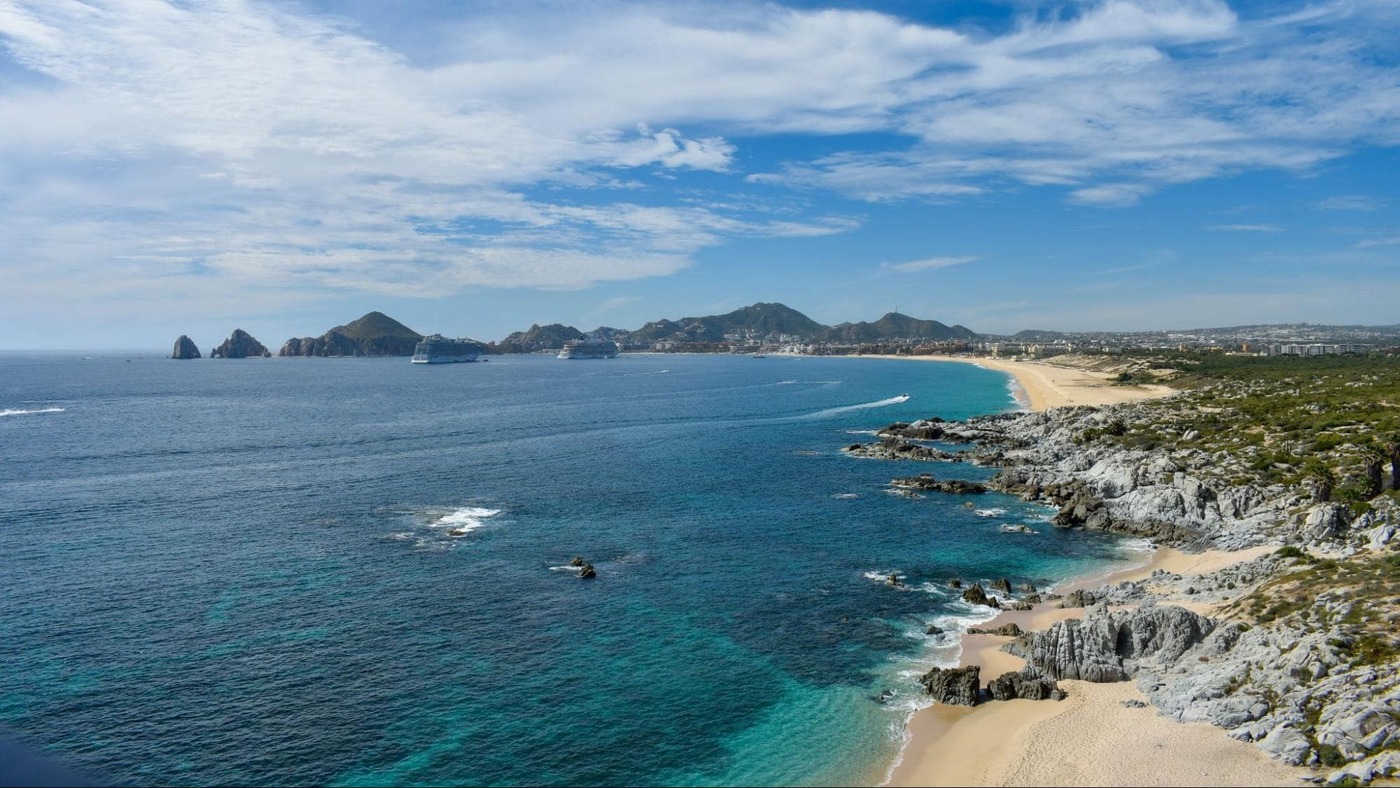 A panoramic view of Cabo San Lucas coastline with turquoise waters, rocky cliffs, sandy beaches, and cruise ships anchored near the famous sea arch.