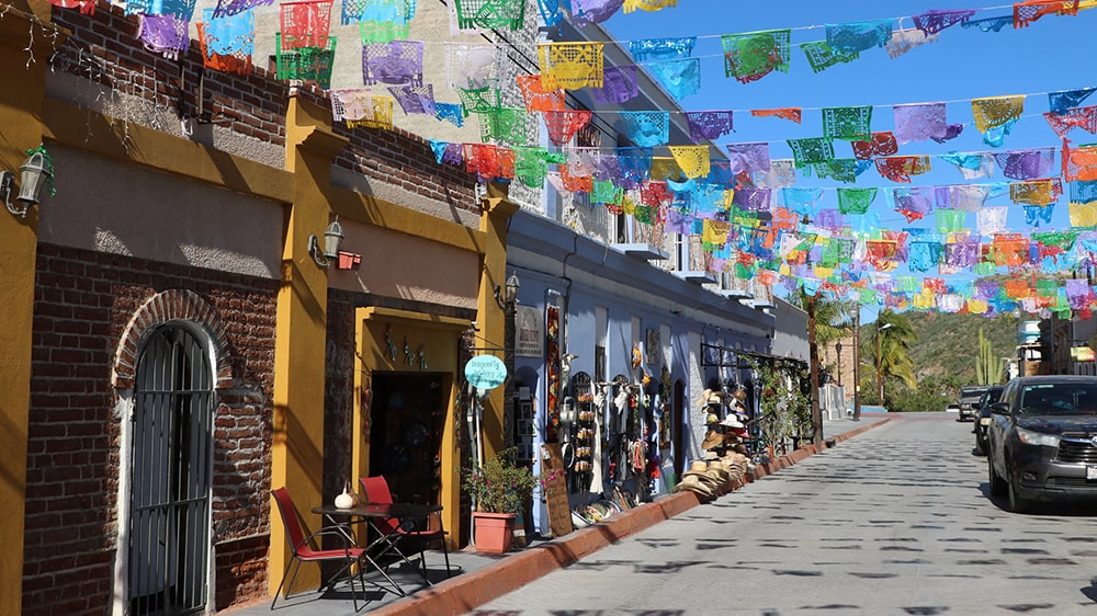 Colorful papel picado flags hanging over a vibrant street lined with shops and cafes in a historic Mexican town.
