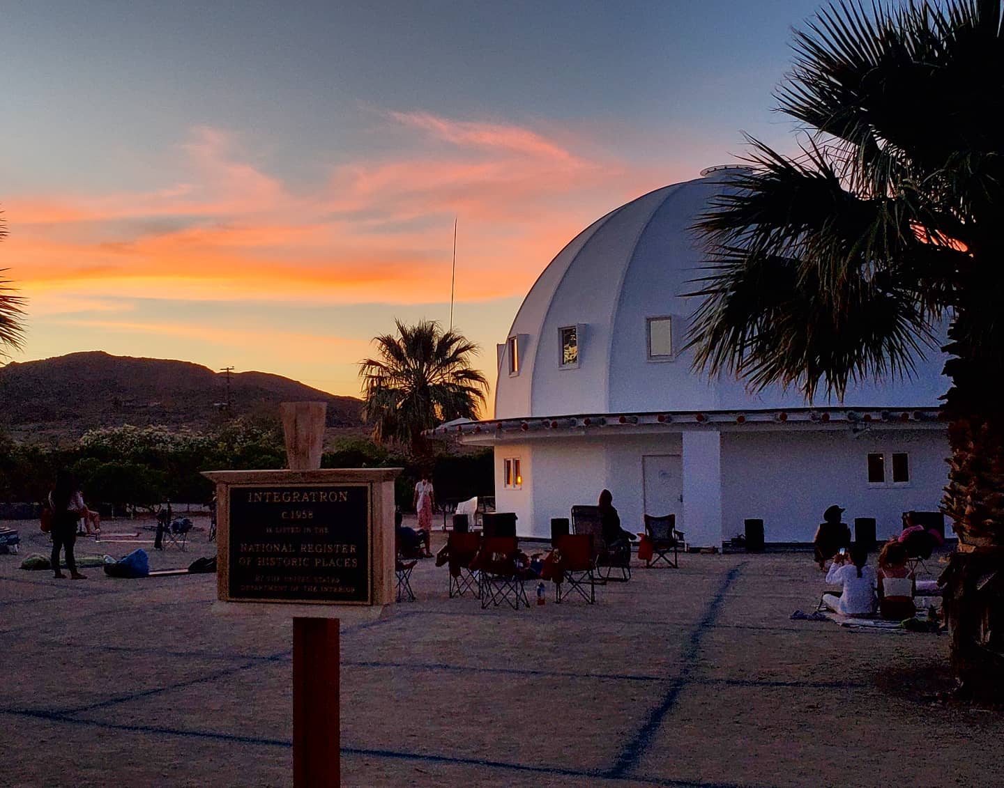 Sunset at the Integratron in Landers, California, with visitors gathered around the iconic white dome surrounded by palm trees and desert mountains, a historic landmark for sound healing.