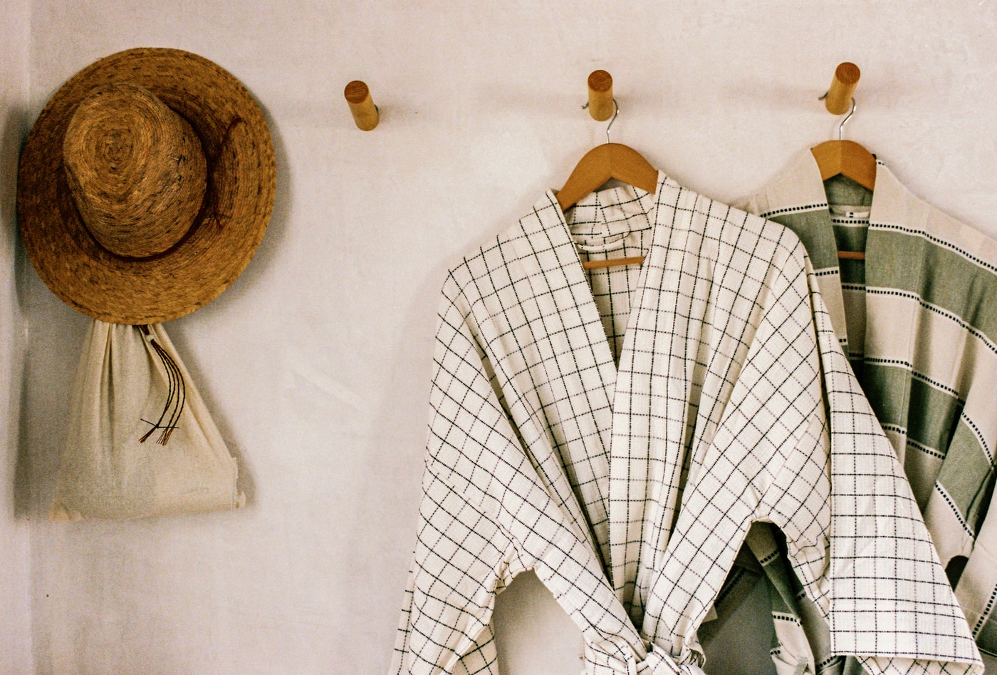 Minimalist home entryway with straw hat, cotton bag, and two checkered linen robes hanging on wooden wall hooks