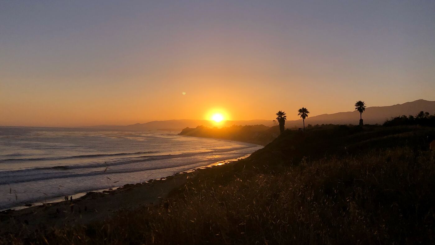 Golden sunset over a peaceful coastal landscape with palm trees, gentle ocean waves, and mountains in the distance