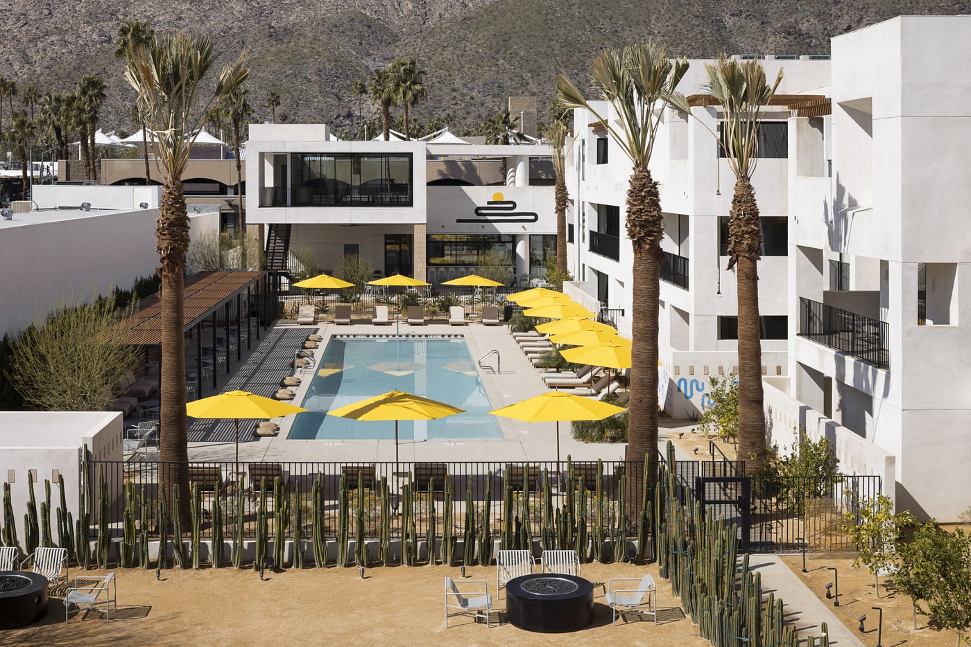 Modern desert hotel courtyard with swimming pool, yellow umbrellas, tall palm trees, and mountain backdrop in Palm Springs
