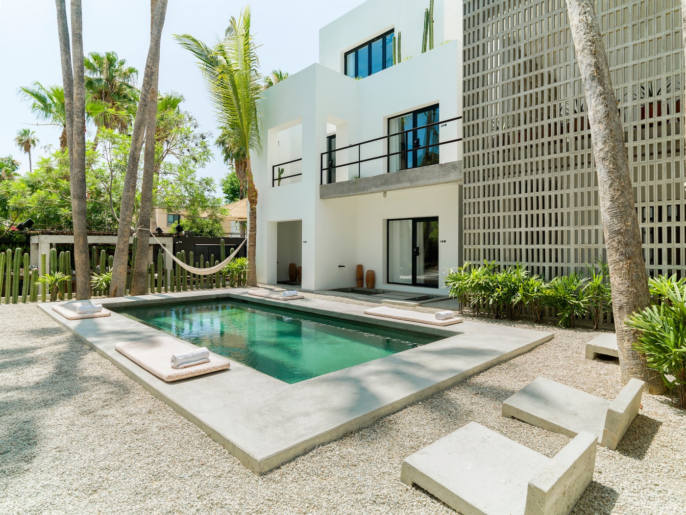 A tranquil outdoor pool area surrounded by lush greenery, modern white architecture, and tall palm trees, with a hammock adding to the relaxing ambiance.